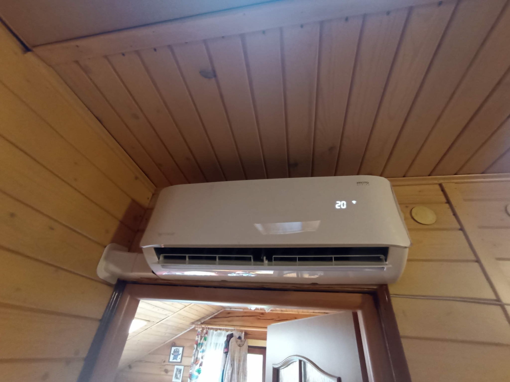 Biała klimatyzacja ścienna z wyświetlaczem 20 stopni, zamontowana w drewnianym wnętrzu. Widok z dołu, fragment otwartego pomieszczenia w tle.