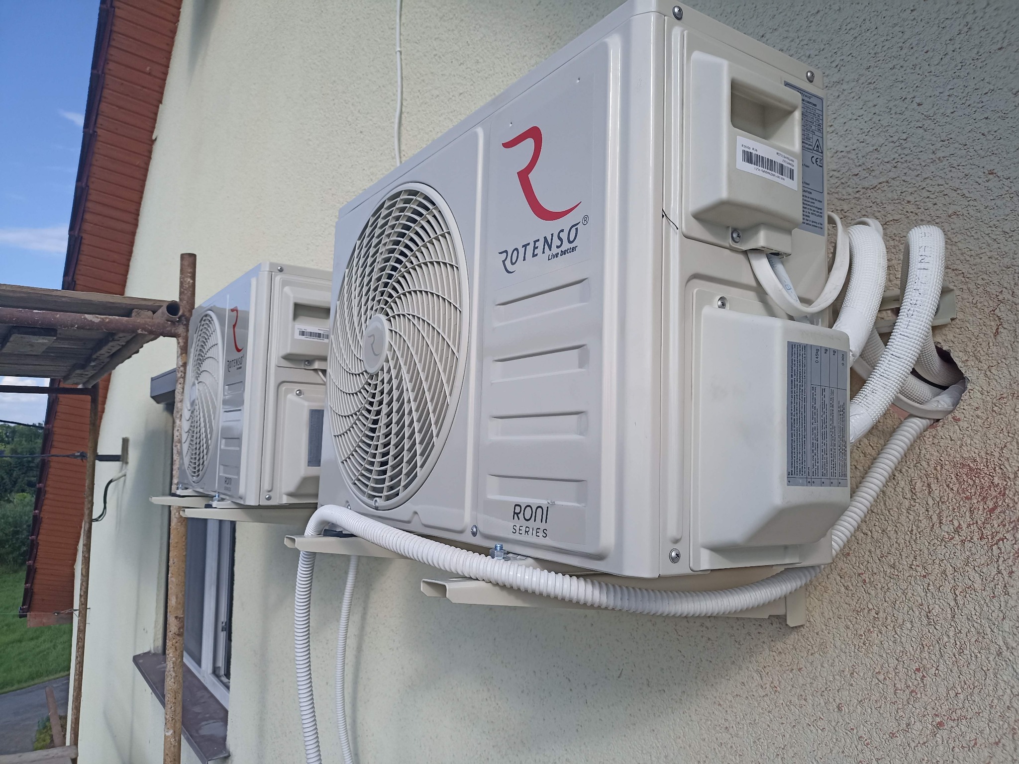 Dwa zamontowane klimatyzatory Rotenso na elewacji budynku, widoczne białe rury instalacyjne i fragment rusztowania po lewej stronie. Ujęcie z dołu.