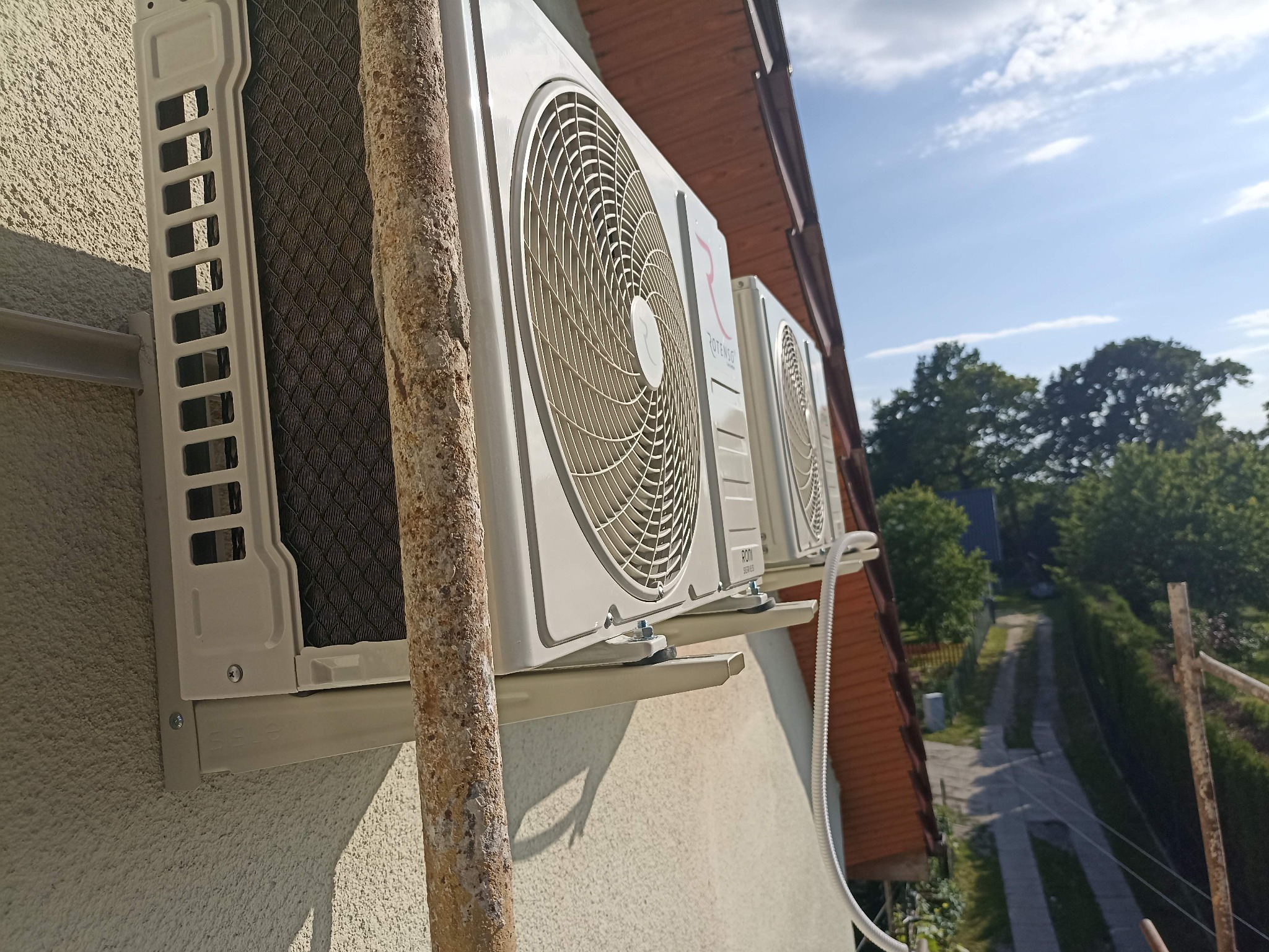 Dwa klimatyzatory Rotenso zamontowane na elewacji budynku, widoczne z boku, z fragmentem rynny i niebem w tle. Instalacja zewnętrzna klimatyzacji.