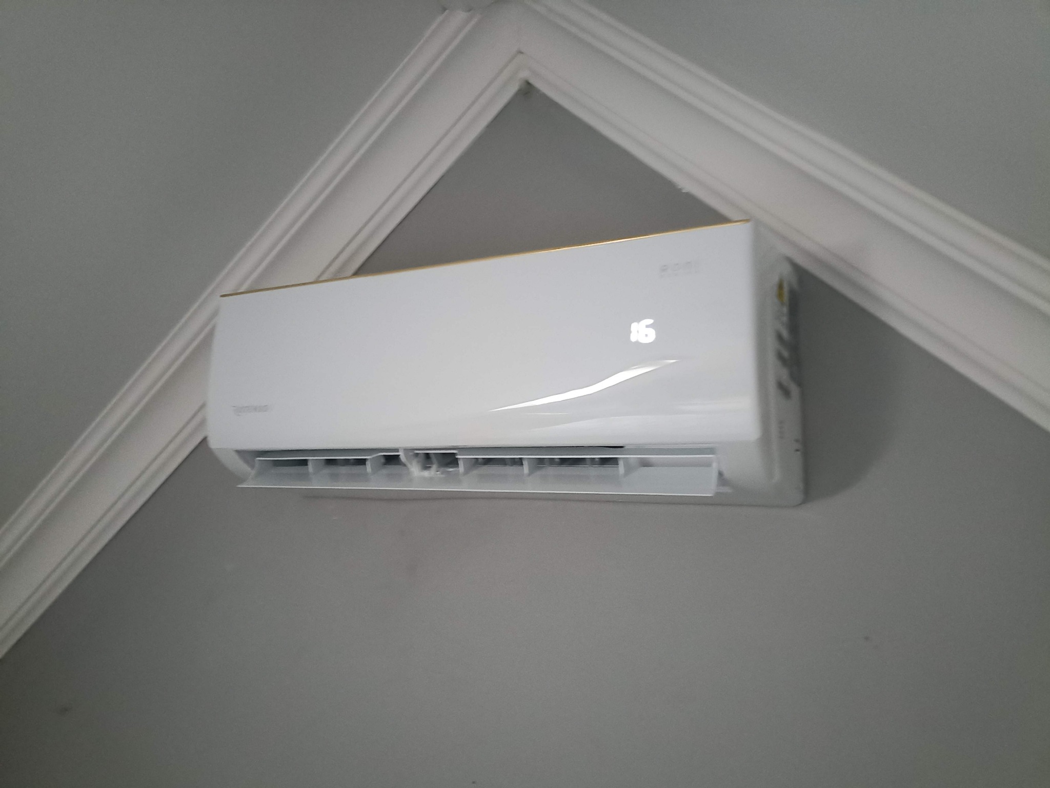Biała klimatyzacja ścienna z wyświetlaczem temperatury, zamontowana w rogu pokoju z dekoracyjną listwą przysufitową. Widok z dołu, lekki kąt.