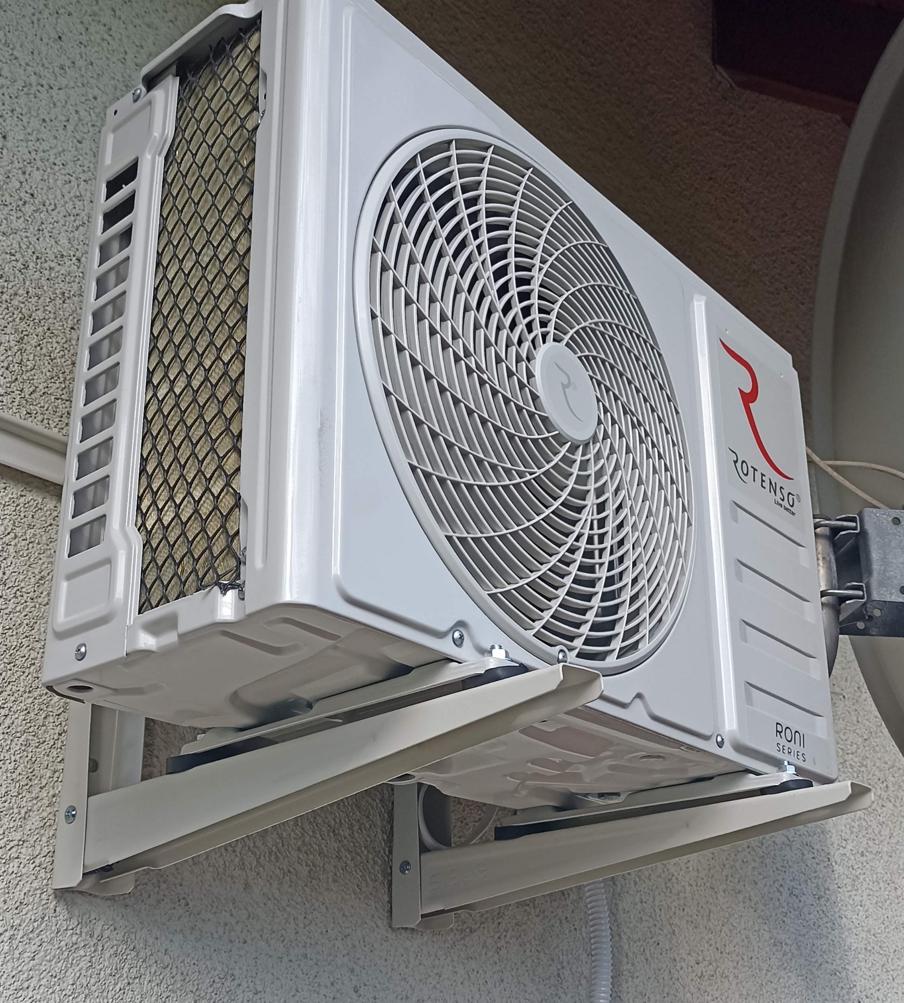 Biała jednostka zewnętrzna klimatyzacji Rotenso Roni Series zamontowana na ścianie budynku, widoczna z boku, z kratką ochronną i logo producenta.