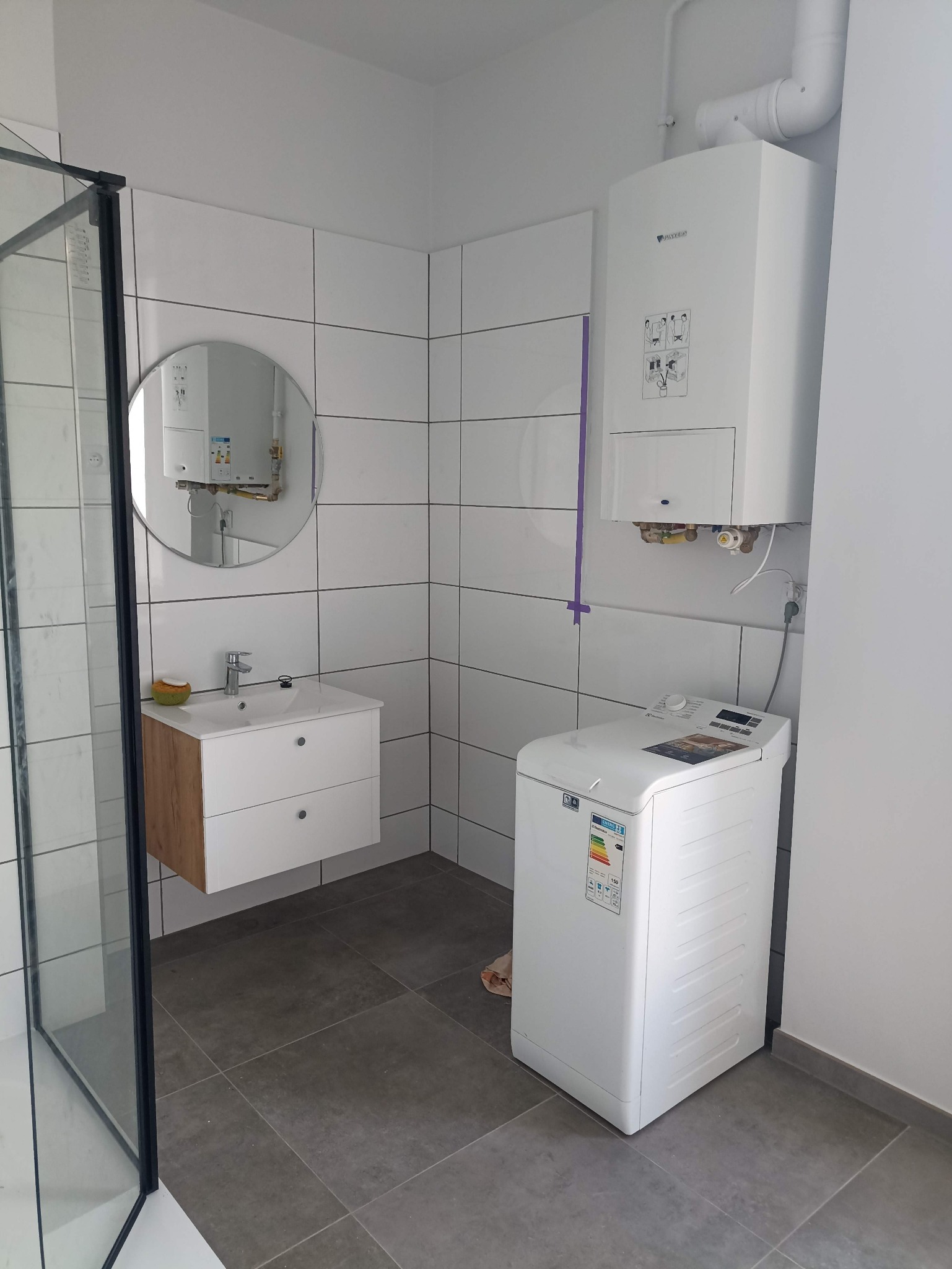 Biała łazienka z piecem gazowym Junkers, pralką Electrolux i umywalką z szafką. Widoczne białe płytki, szara podłoga i kabina prysznicowa z czarnymi ramami.