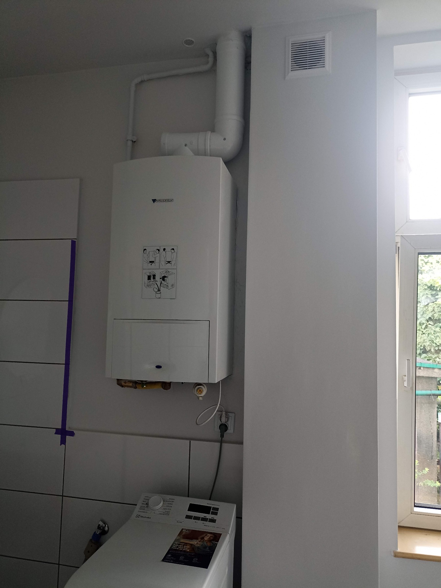 Biały piec gazowy Junkers zamontowany na ścianie w łazience, widoczne rury odprowadzające spaliny, pralka Electrolux w dolnej części kadru, okno z widokiem na zieleń.