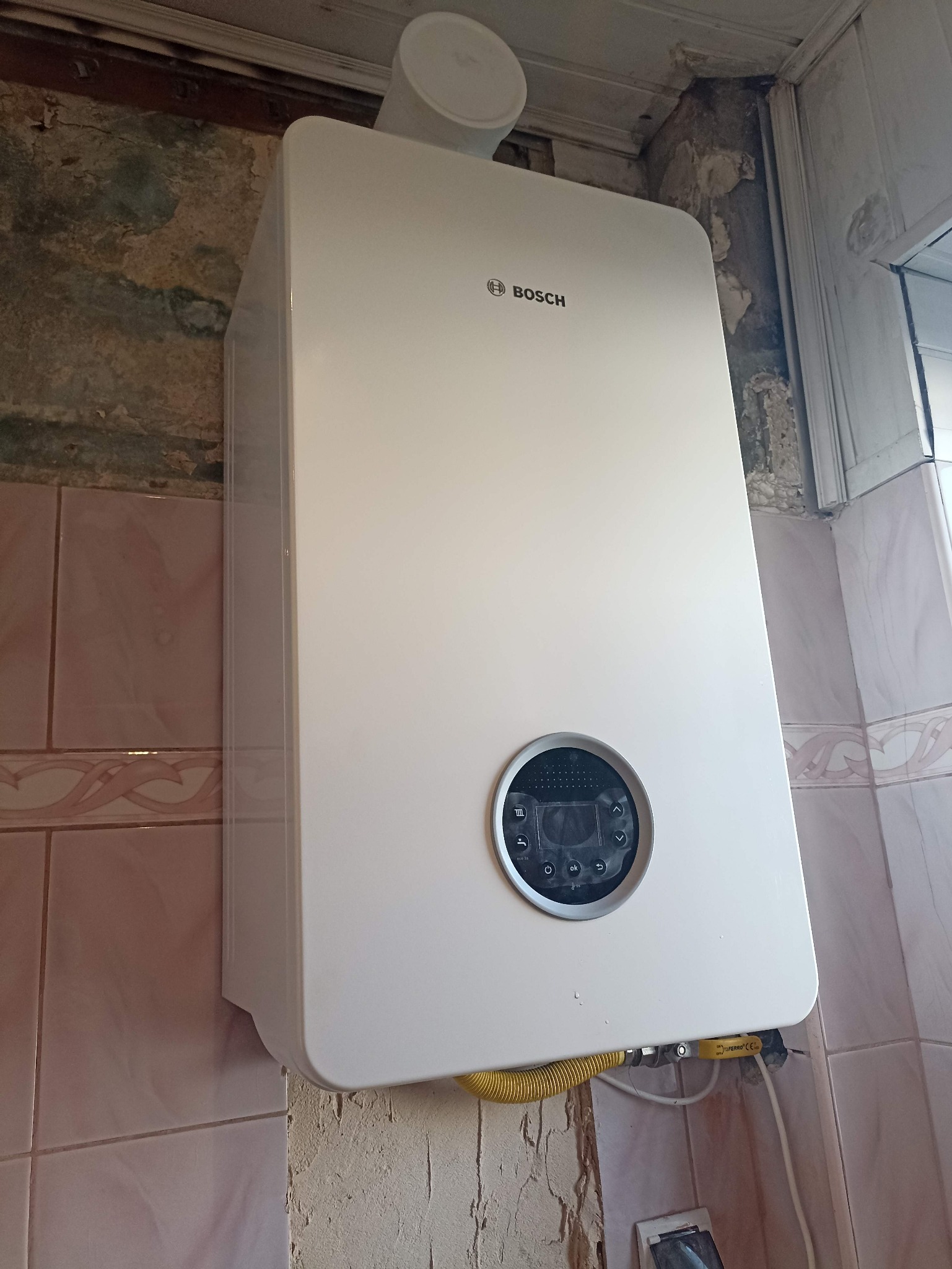 Biały, wiszący piec gazowy marki Bosch z żółtym przewodem gazowym, zamontowany na tle ściany z różowymi płytkami i fragmentem surowego muru.