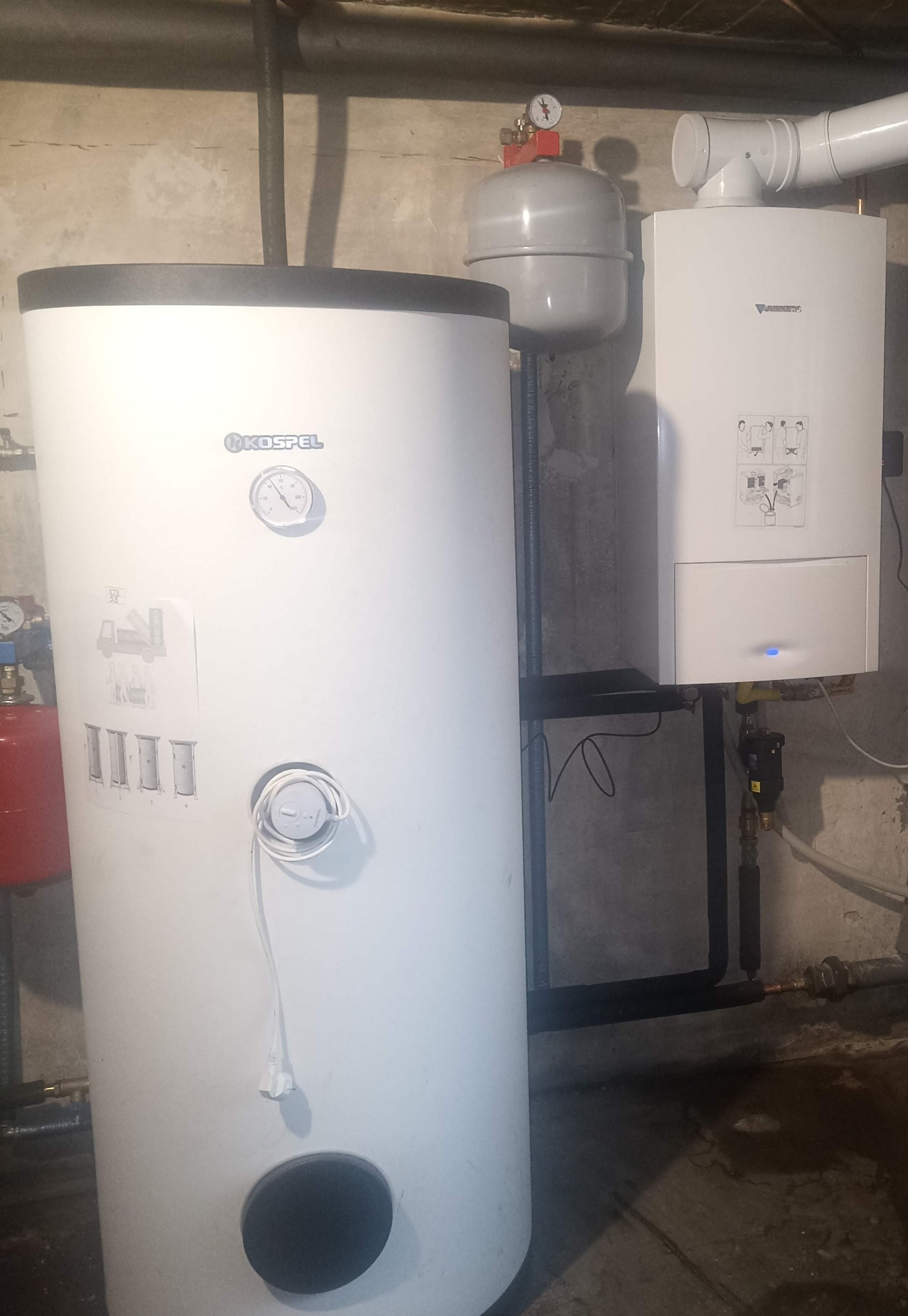Biały piec gazowy Junkers i zasobnik Kospel w kotłowni. Widoczne rury, manometry i instrukcje obsługi na urządzeniach. Surowe ściany w tle.