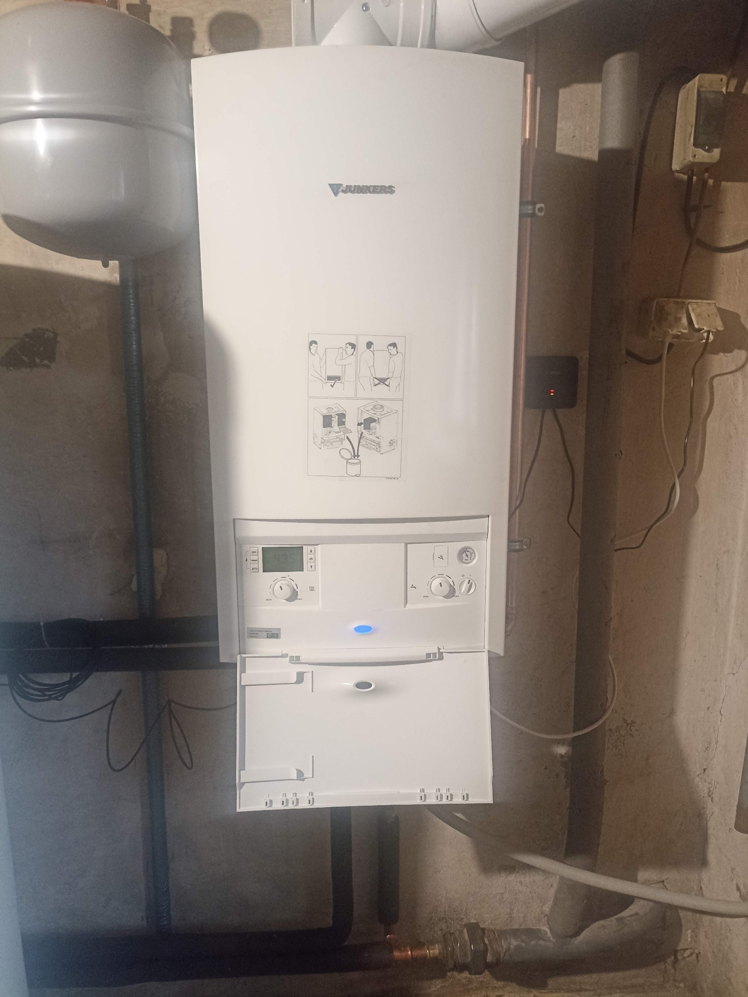 Biały, wiszący piec gazowy Junkers na tle ściany z widocznymi rurami. Otwarta dolna klapka pieca, widoczne elementy sterujące i schemat montażu na obudowie.
