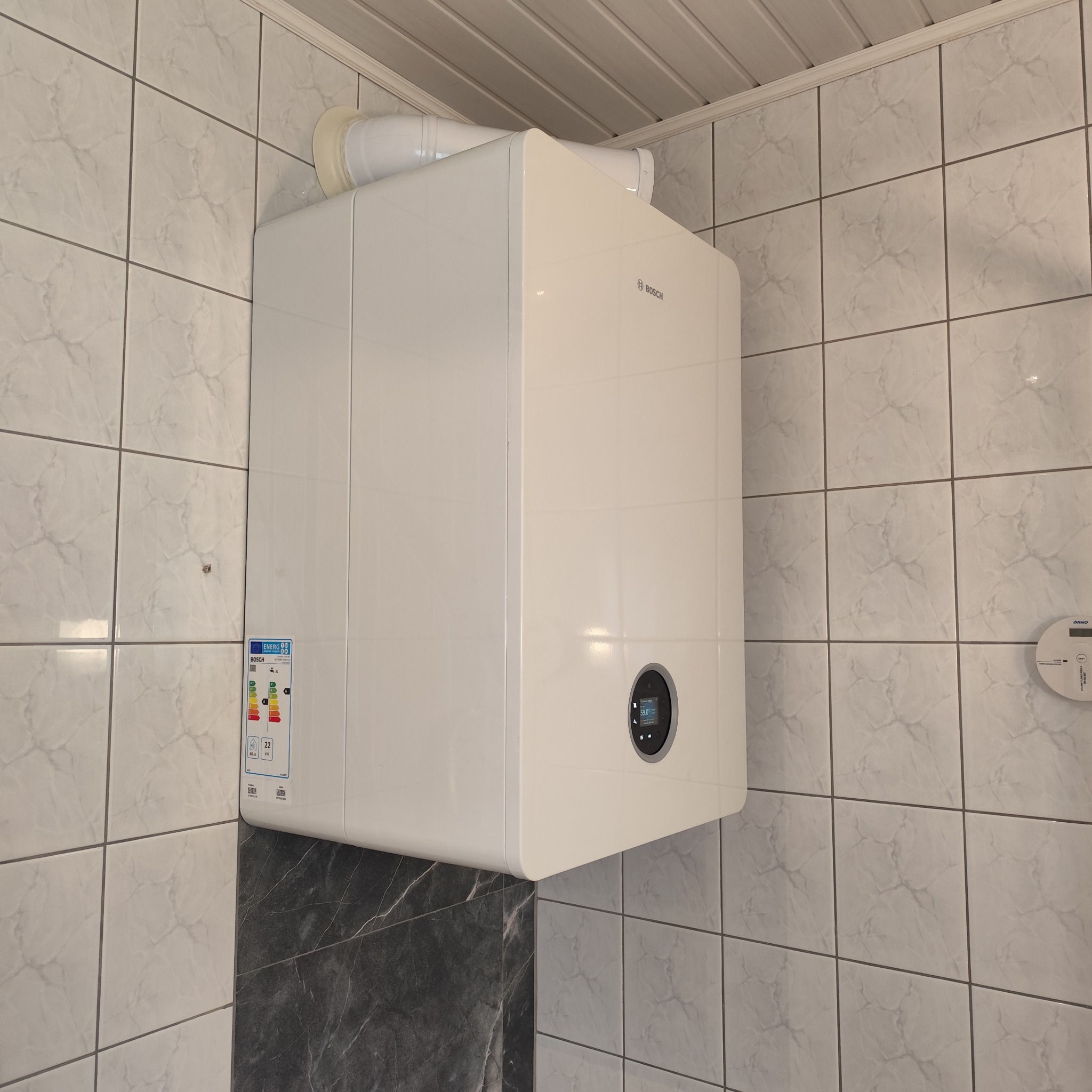 Biały, nowoczesny piec gazowy Bosch zamontowany na ścianie z płytek, z widoczną etykietą energetyczną i elementami instalacji. Ujęcie z dołu, w rogu pomieszczenia.