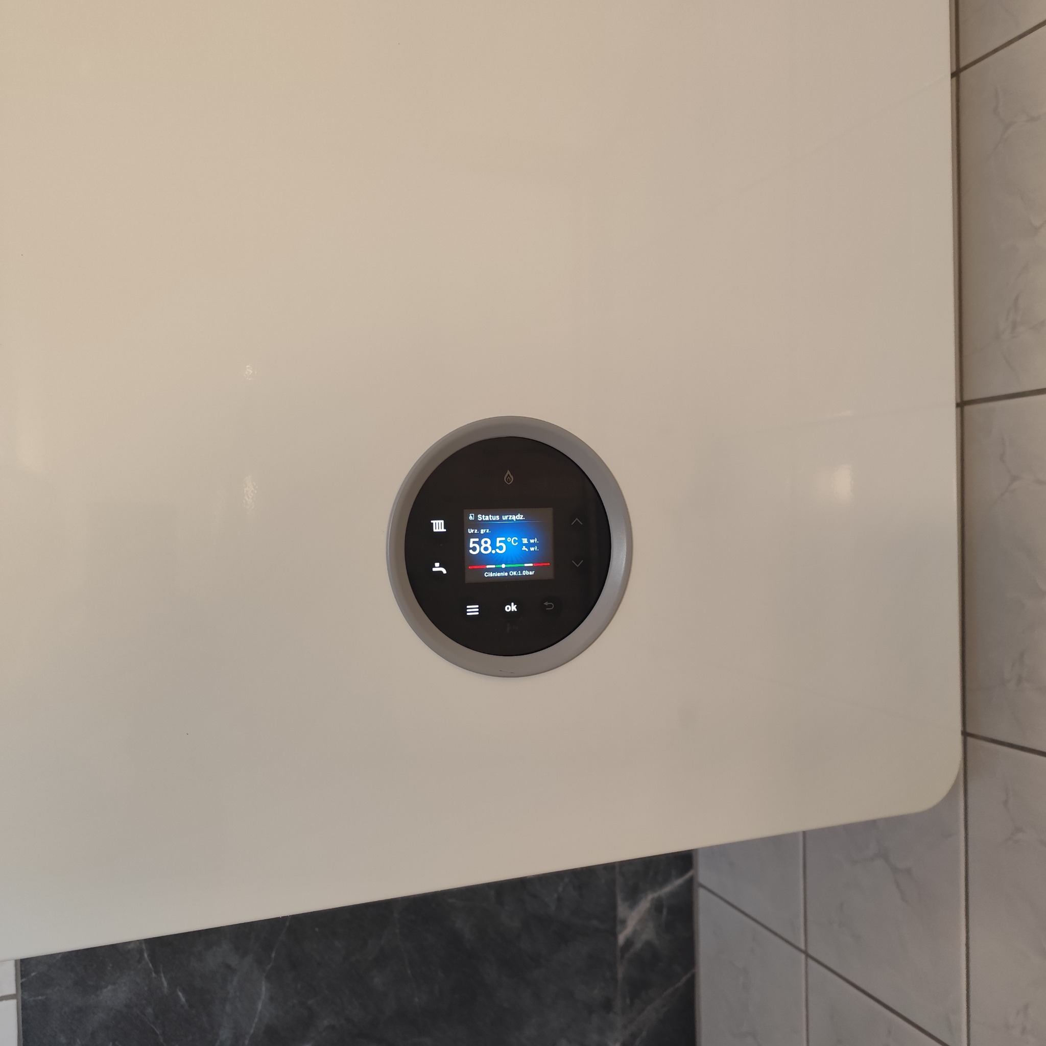 Nowoczesny piec gazowy z wyświetlaczem wskazującym temperaturę 58.5°C i ciśnienie 1.0 bar, zamontowany na tle płytek imitujących marmur.