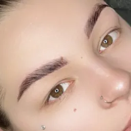 Zbliżenie na brwi po laminacji, widoczne pojedyncze włoski, piercing w nosie i na policzku, brązowe oczy z refleksami światła.