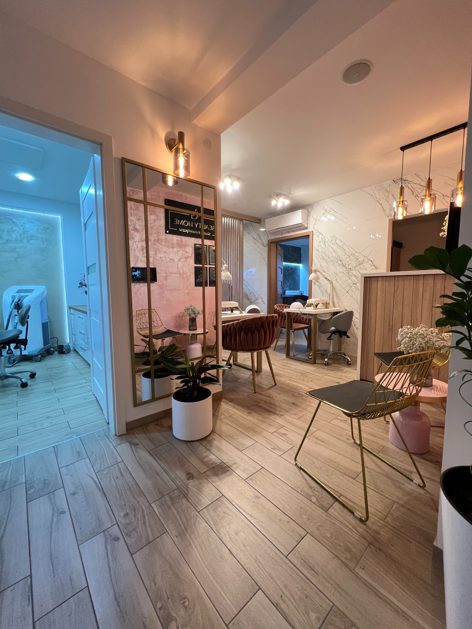 Jasne wnętrze salonu z widocznymi stanowiskami do manicure i pedicure, lustrem z napisem 'Beauty Home' i roślinami w doniczkach.