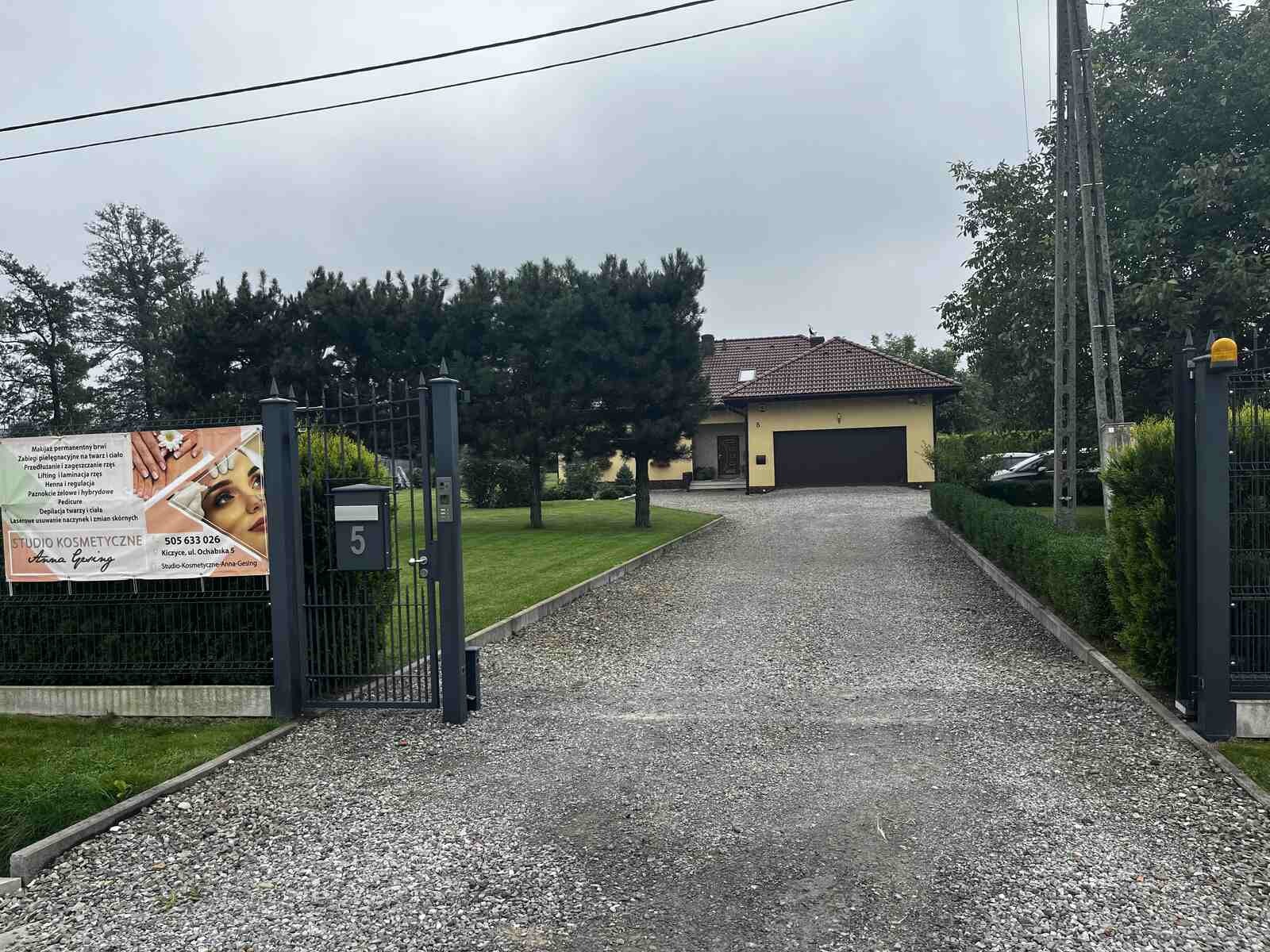 Wjazd do salonu kosmetycznego Anna Gesing z reklamą usług na bramie: manicure, makijaż permanentny, depilacja laserowa i inne zabiegi.