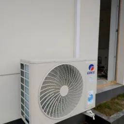 Zewnętrzna jednostka klimatyzacji marki Gree zamontowana na elewacji budynku, widoczny fragment ściany i otwarte drzwi do wnętrza w stanie surowym.