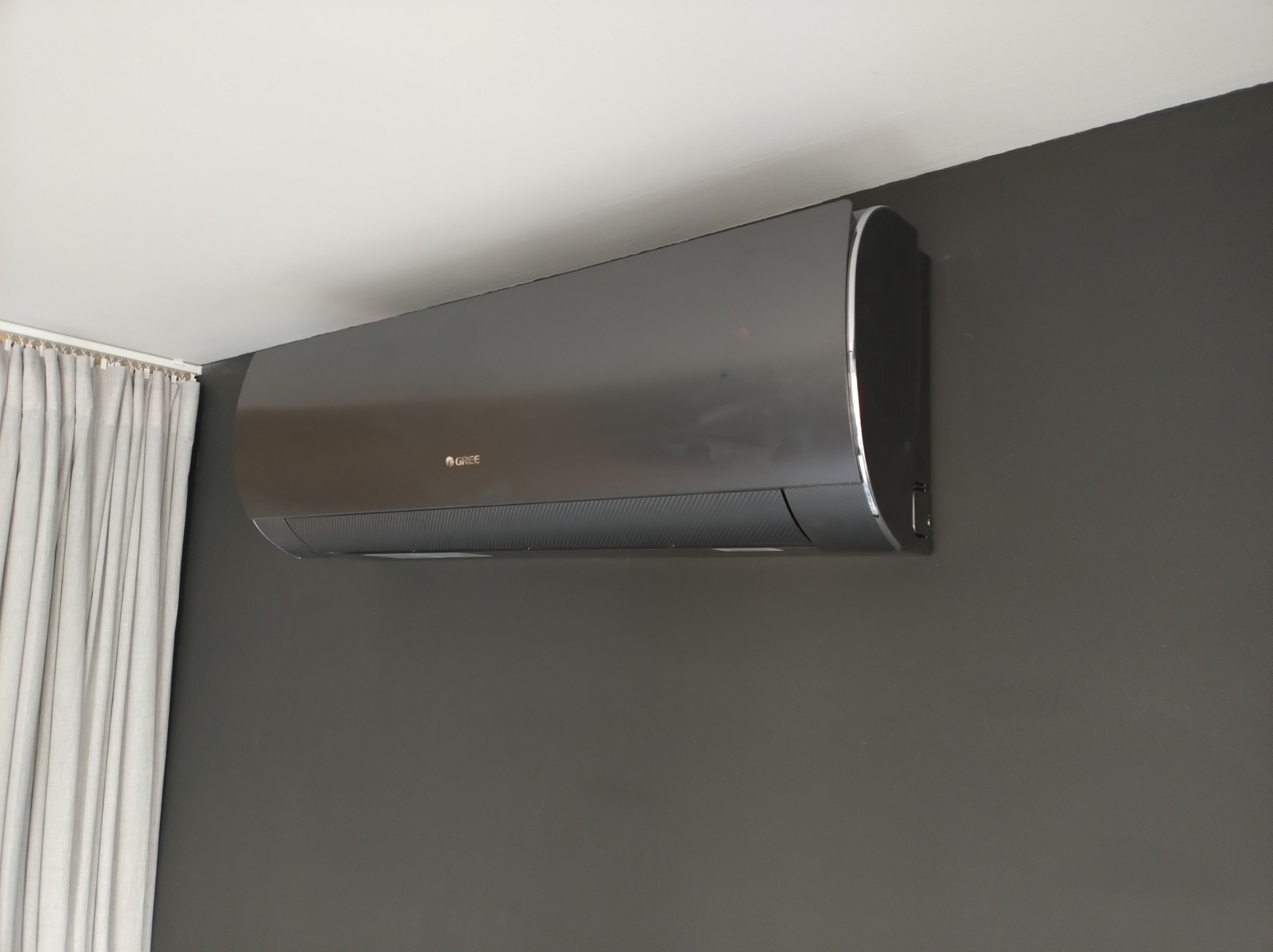 Szary klimatyzator marki Gree zamontowany na ciemnoszarej ścianie, widoczne białe zasłony po lewej stronie.