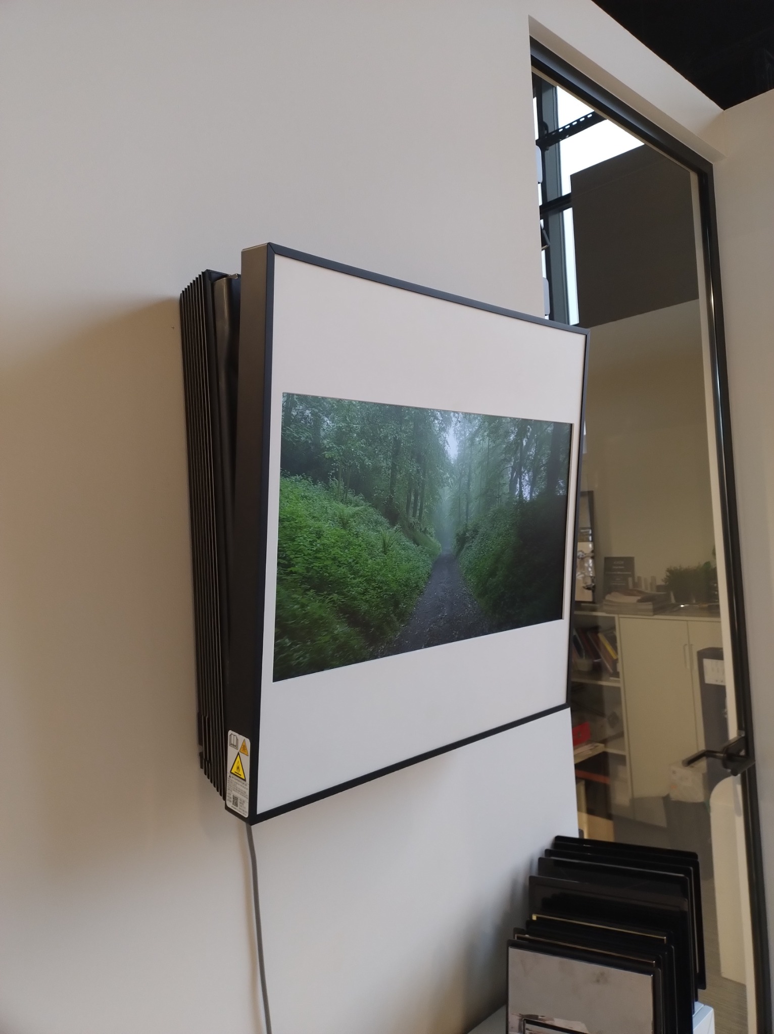 Wiszący na ścianie rekuperator z wbudowanym ekranem wyświetlającym leśny krajobraz, widoczne oznaczenia bezpieczeństwa na obudowie i kabel zasilający.