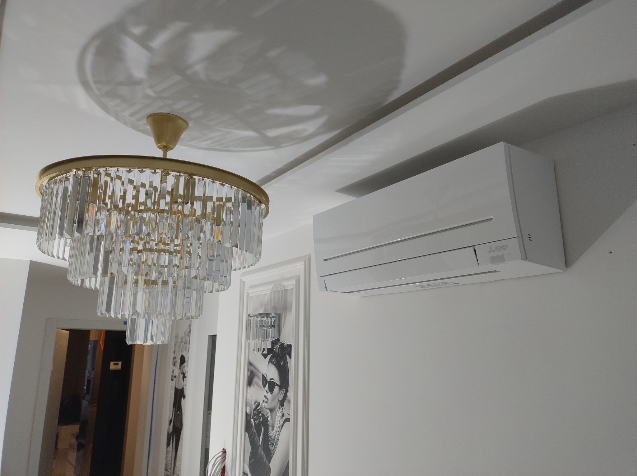 Biała jednostka klimatyzacji Mitsubishi Electric zamontowana na ścianie w eleganckim wnętrzu z kryształowym żyrandolem i dekoracyjnym obrazem.
