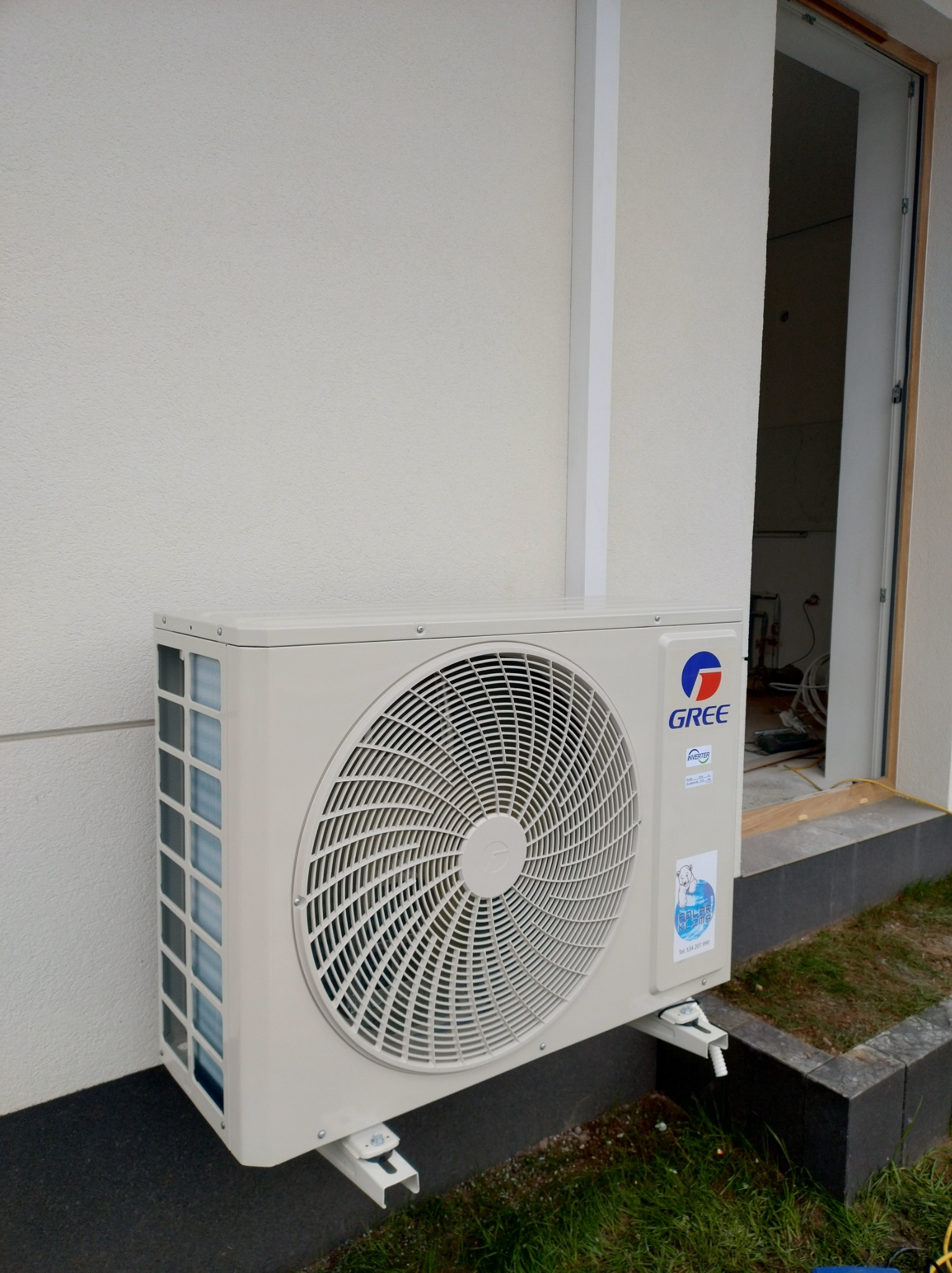 Zewnętrzna jednostka klimatyzacji marki Gree zamontowana na elewacji budynku, widoczny fragment ściany i otwarte drzwi do wnętrza w stanie surowym.