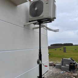 NR ONE RAFAŁ NOWICKI - Zewnętrzna jednostka klimatyzacji Rotenso na podnośniku teleskopowym podczas montażu przy ścianie budynku, widoczne przewody instalacyjne i narzędzia w tle.
