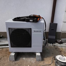 NR ONE RAFAŁ NOWICKI - Biała pompa ciepła marki Buderus, zamontowana na betonowych podstawach przed jasną ścianą budynku, z narzędziami w otwartym pudełku na górze i zwiniętym kablem zasilającym obok.