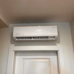 Biały klimatyzator Rotenso Roni Series zamontowany nad drzwiami, wyświetlający temperaturę 18 stopni Celsjusza, w pomieszczeniu o jasnych ścianach.