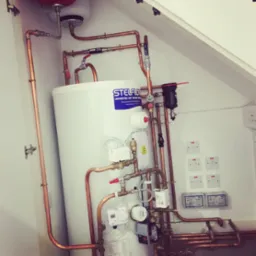 Biały zasobnik ciepłej wody użytkowej StelFlow z licznymi miedzianymi rurami, zaworami i elementami sterującymi zamontowany w pomieszczeniu z białymi ścianami i włącznikami światła.