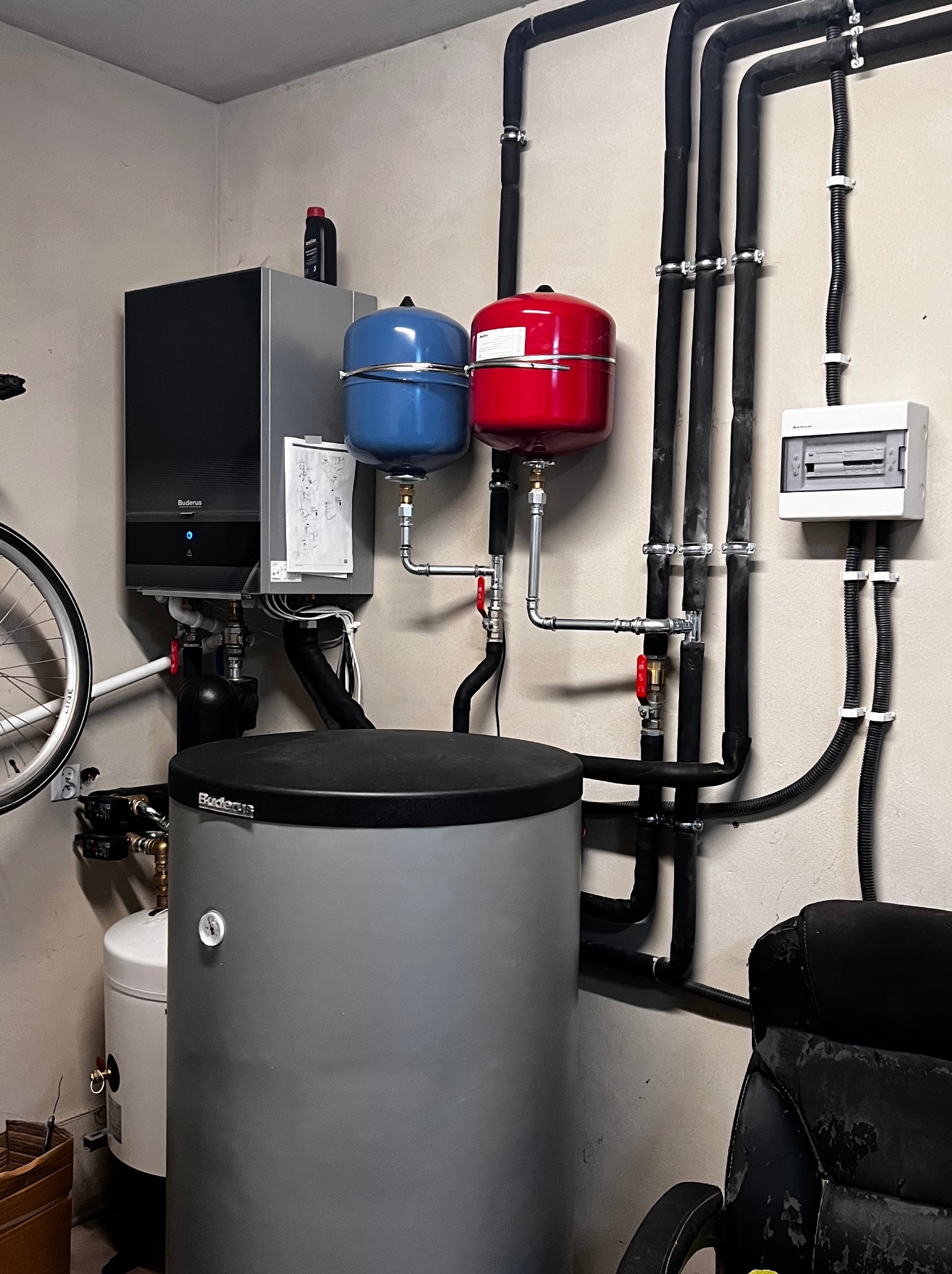 Nowoczesny piec gazowy Buderus z zasobnikiem, naczyniami przeponowymi w kolorze czerwonym i niebieskim oraz rozbudowaną instalacją hydrauliczną z czarnych rur, zamontowany w pomieszczeniu...