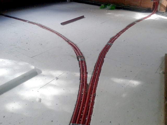 Czerwone rury grzewcze ułożone w rowkach w izolacji podłogowej z białego styropianu, rozchodzące się w dwie strony.