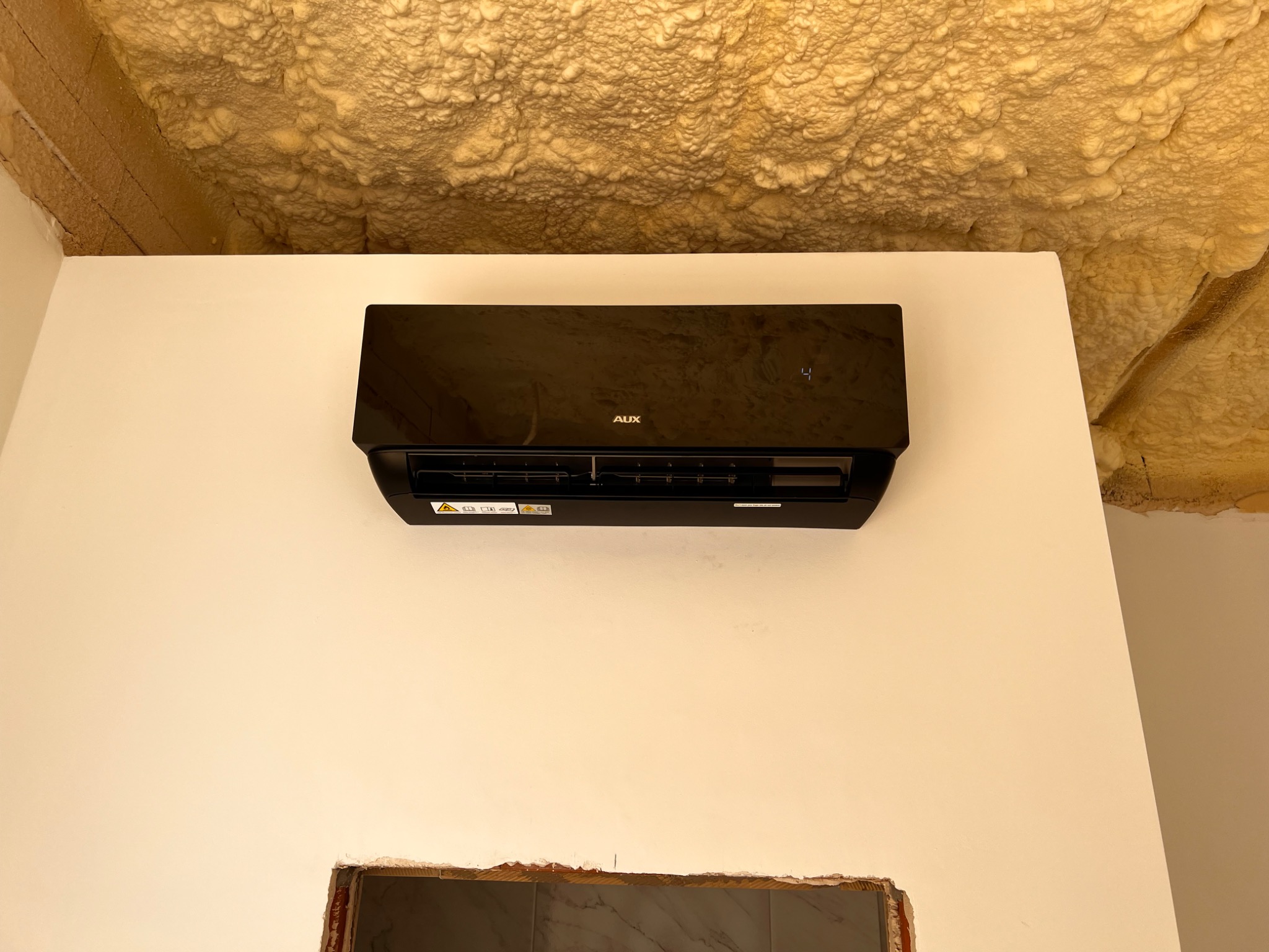Czarny klimatyzator AUX zamontowany na białej ścianie, z widoczną izolacją piankową na suficie i niedokończonym otworem w ścianie poniżej.