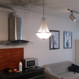 HandyManWaw Artur Nagraba - Salon z szarą sofą, komodą z mikrofalówką, okapem kuchennym i kryształowym żyrandolem wiszącym pod sufitem z plamami.