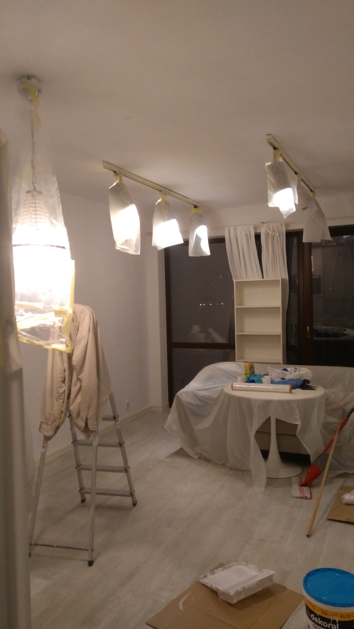 Pokój przygotowany do malowania: meble i lampy zabezpieczone folią, drabina oparta o ścianę, widoczne wiadro z farbą i kuweta malarska na podłodze.
