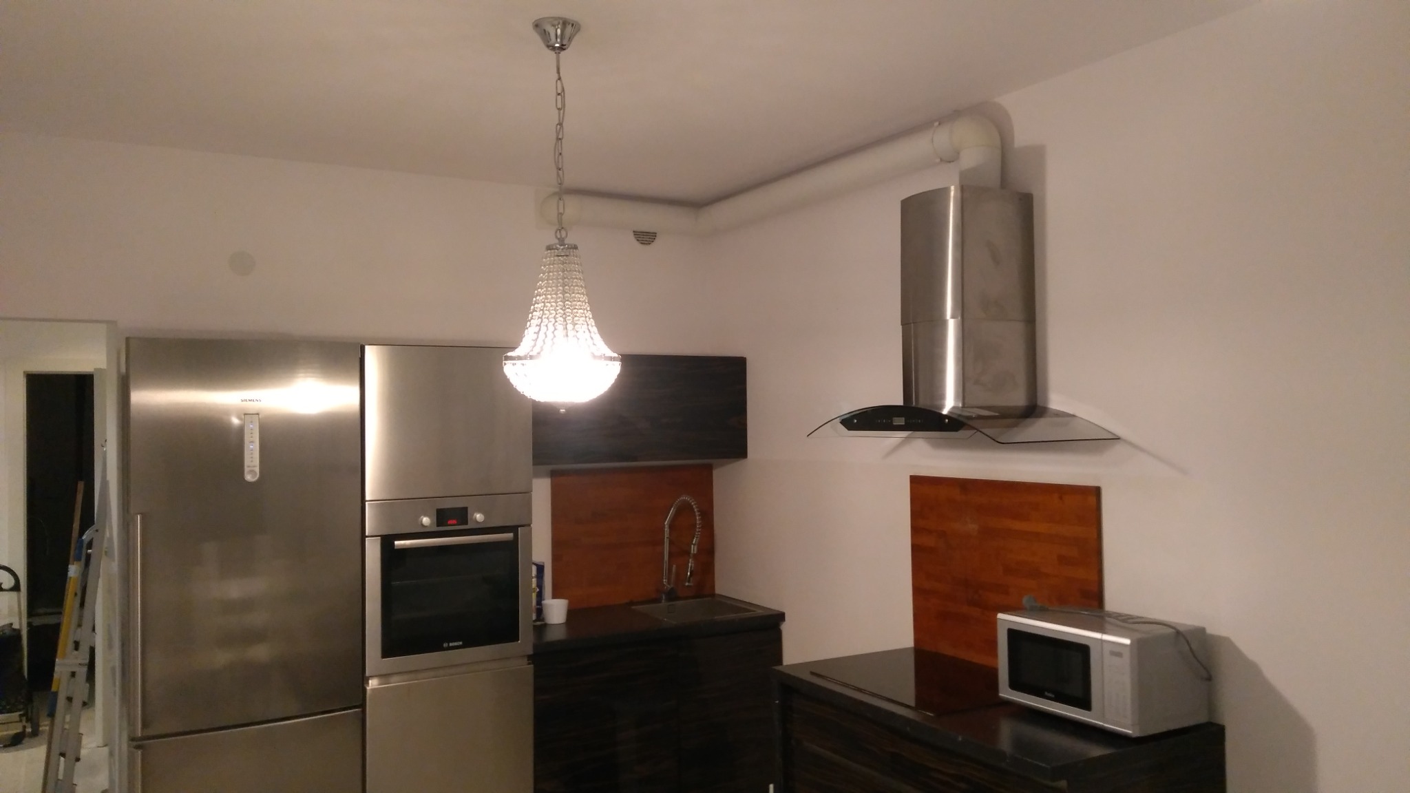 Nowoczesna kuchnia z zabudową w odcieniach szarości i brązu, oświetlona kryształowym żyrandolem, z widocznym sprzętem AGD: lodówka, piekarnik, mikrofalówka i okap.