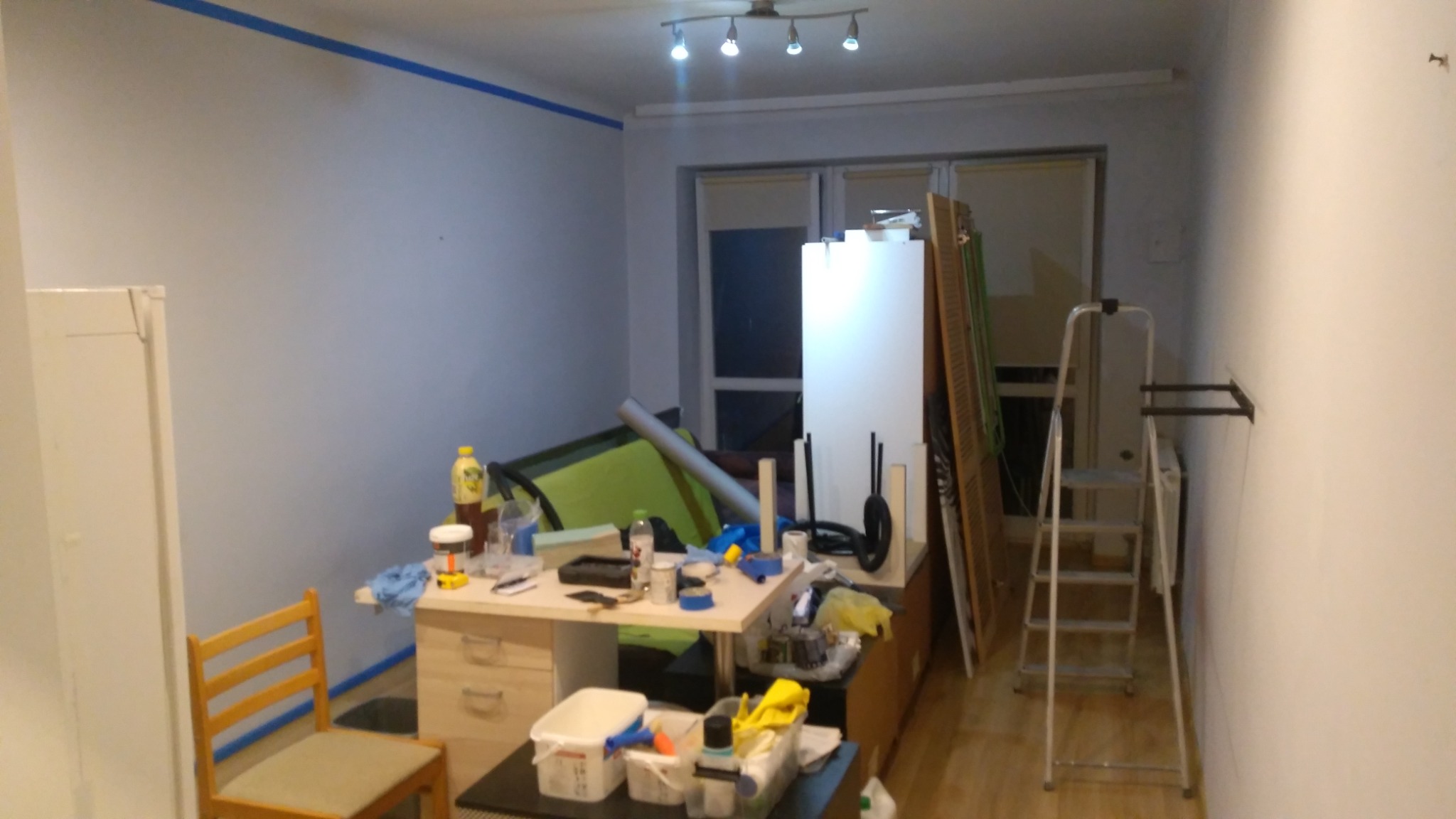 Pokój w trakcie przygotowań do prac wykończeniowych: ściany zabezpieczone taśmą malarską, widoczne narzędzia, drabina, meble przesunięte na środek pomieszczenia, oświetlenie sufitowe.