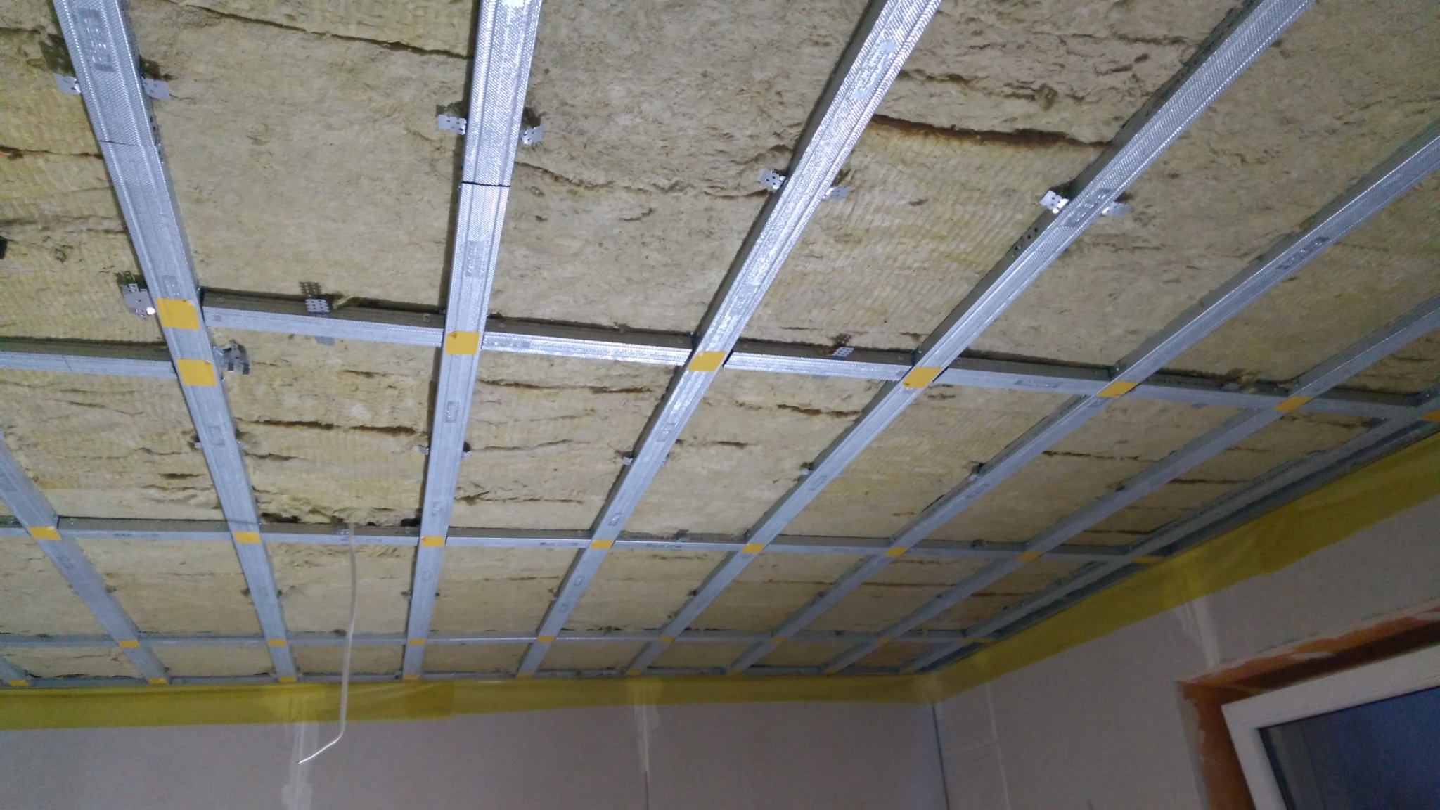 Sufit podwieszany w trakcie montażu: widoczna konstrukcja metalowa z izolacją z wełny mineralnej, zabezpieczona żółtą folią malarską przy ścianach.