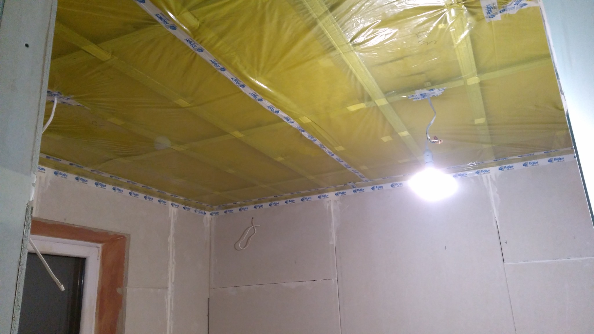 Sufit podwieszany w trakcie montażu, widoczna żółta folia izolacyjna, profile Rigips na ścianach i suficie, tymczasowe oświetlenie żarówką