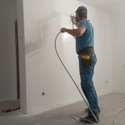 Usługi remontowo budowlane - Pracownik w masce i kombinezonie maluje ścianę na biało za pomocą pistoletu natryskowego. Widoczne przewody i narzędzia malarskie na podłodze w jasnym wnętrzu.