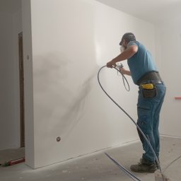 Usługi remontowo budowlane - Malowanie natryskowe ściany wewnątrz pomieszczenia przez pracownika w masce, widoczne narzędzia i detale wykończenia. Ściana świeżo pomalowana na biało.