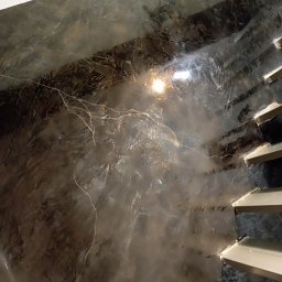 Usługi remontowo budowlane - Ściana z efektem dekoracyjnym w odcieniach szarości i brązu, przypominająca marmur, oświetlona punktowo, widoczne schody w tle. Unikalna faktura, nieregularne wzory.