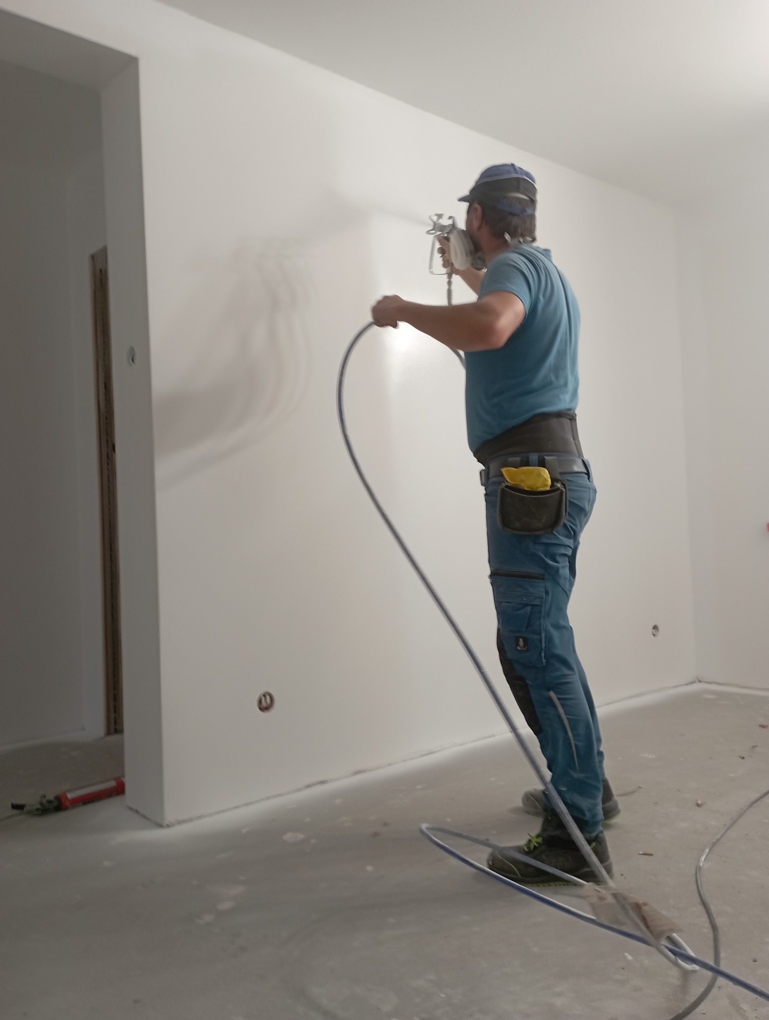 Pracownik w masce i kombinezonie maluje ścianę na biało za pomocą pistoletu natryskowego. Widoczne przewody i narzędzia malarskie na podłodze w jasnym wnętrzu.