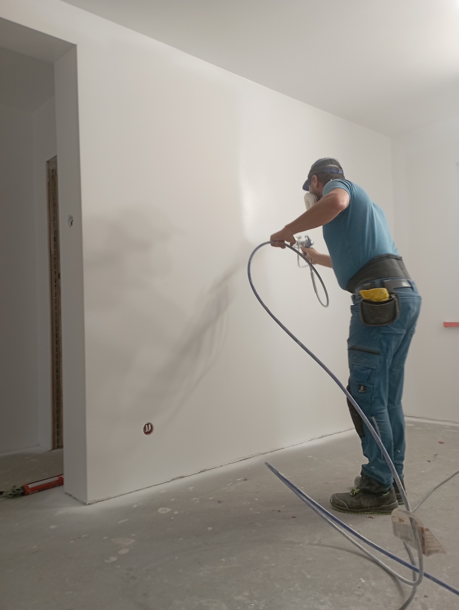 Malowanie natryskowe ściany wewnątrz pomieszczenia przez pracownika w masce, widoczne narzędzia i detale wykończenia. Ściana świeżo pomalowana na biało.