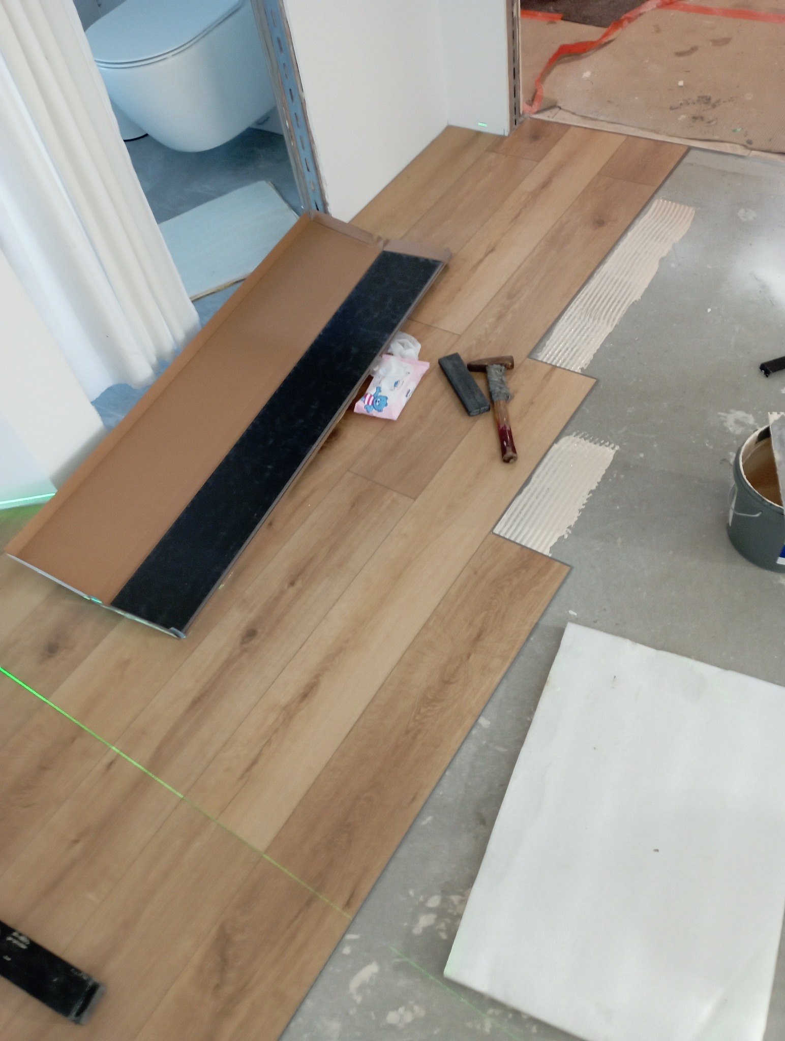 Układanie paneli winylowych w łazience, widoczny laser, młotek, klej i panele o wzorze drewna. Częściowo ułożona podłoga, etap montażu.