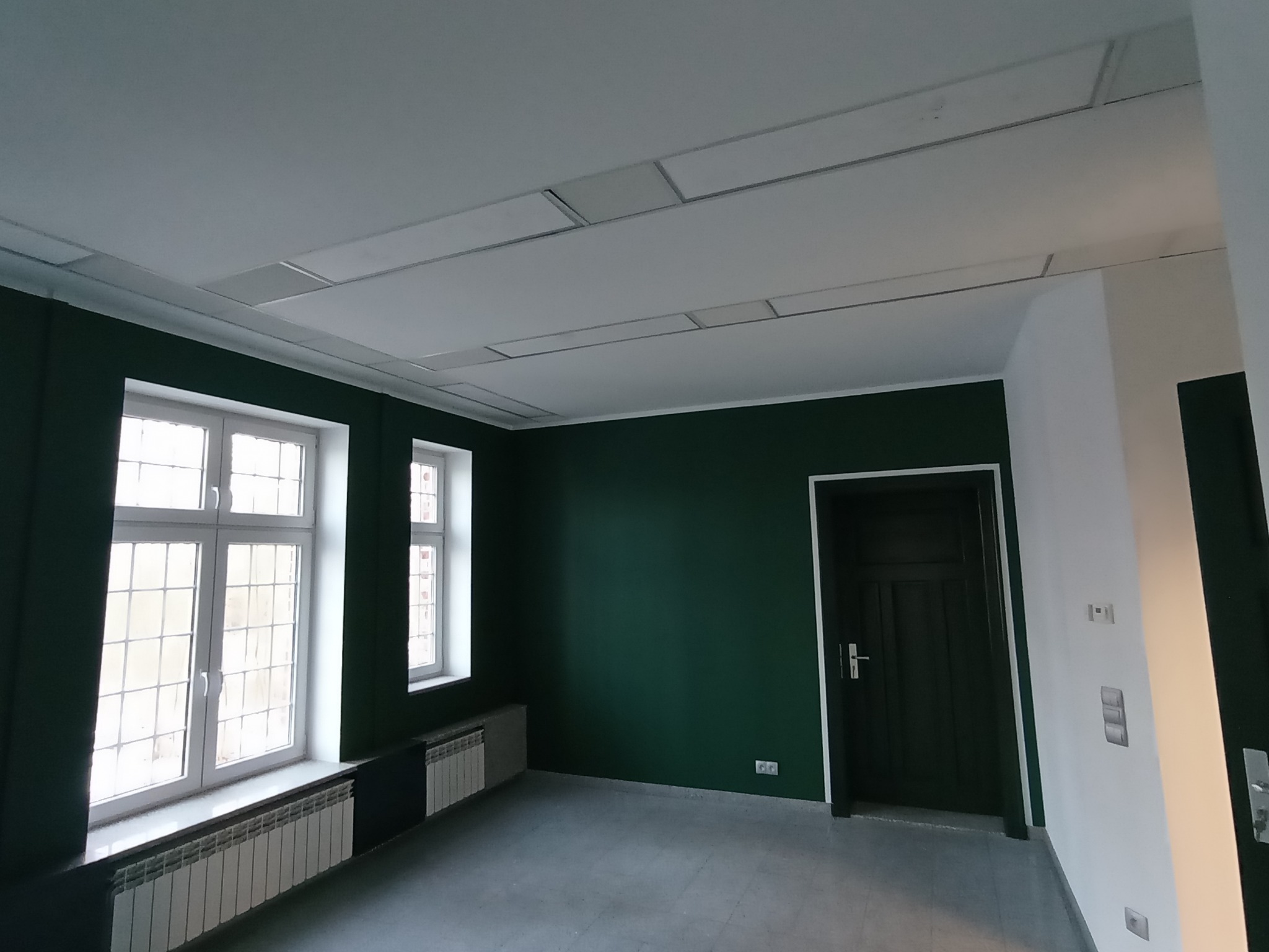 Wnętrze po remoncie: ściany pomalowane na butelkową zieleń, biały sufit z panelami, dwa okna z białymi ramami, grzejniki i ciemne drzwi. Widok z perspektywy podłogi.