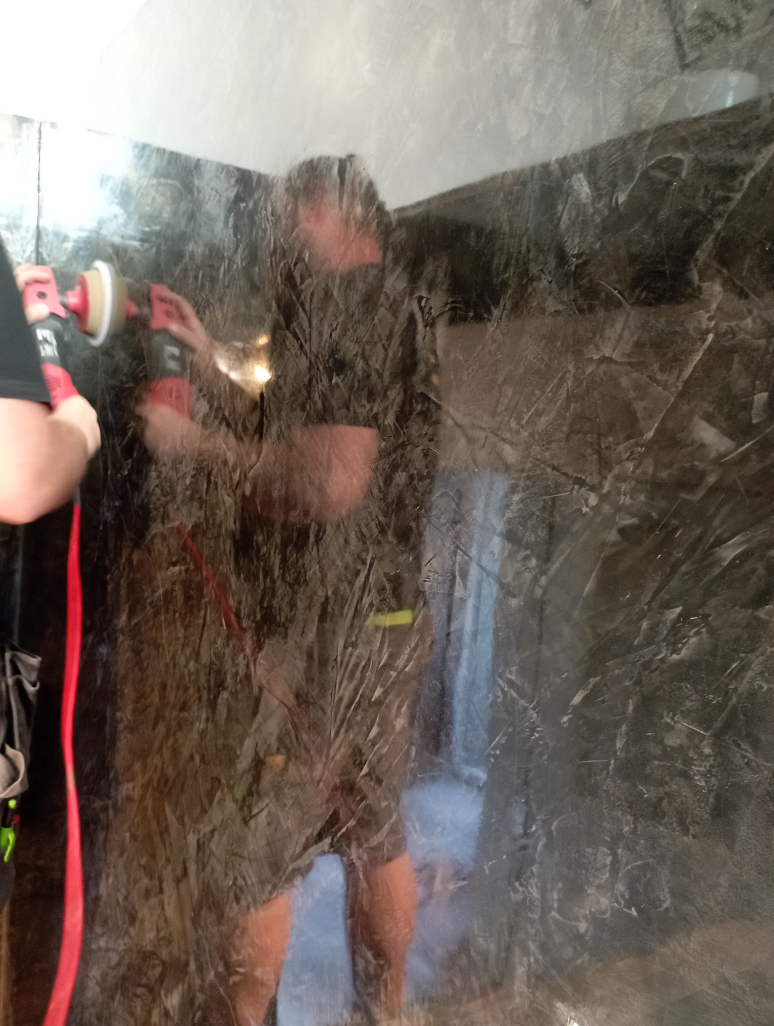 Nakładanie stiuku w odcieniach szarości na ścianę za pomocą polerki. Widoczny fragment ściany z wyraźną fakturą i odbicie pracownika w polerowanej powierzchni.