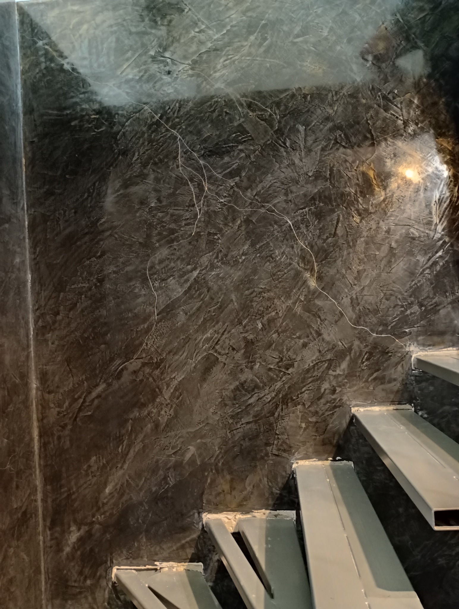 Ściana w stylu betonu architektonicznego w ciemnych odcieniach szarości, z widocznymi schodami. Nowoczesne wykończenie wnętrza z efektem surowości i minimalizmu.