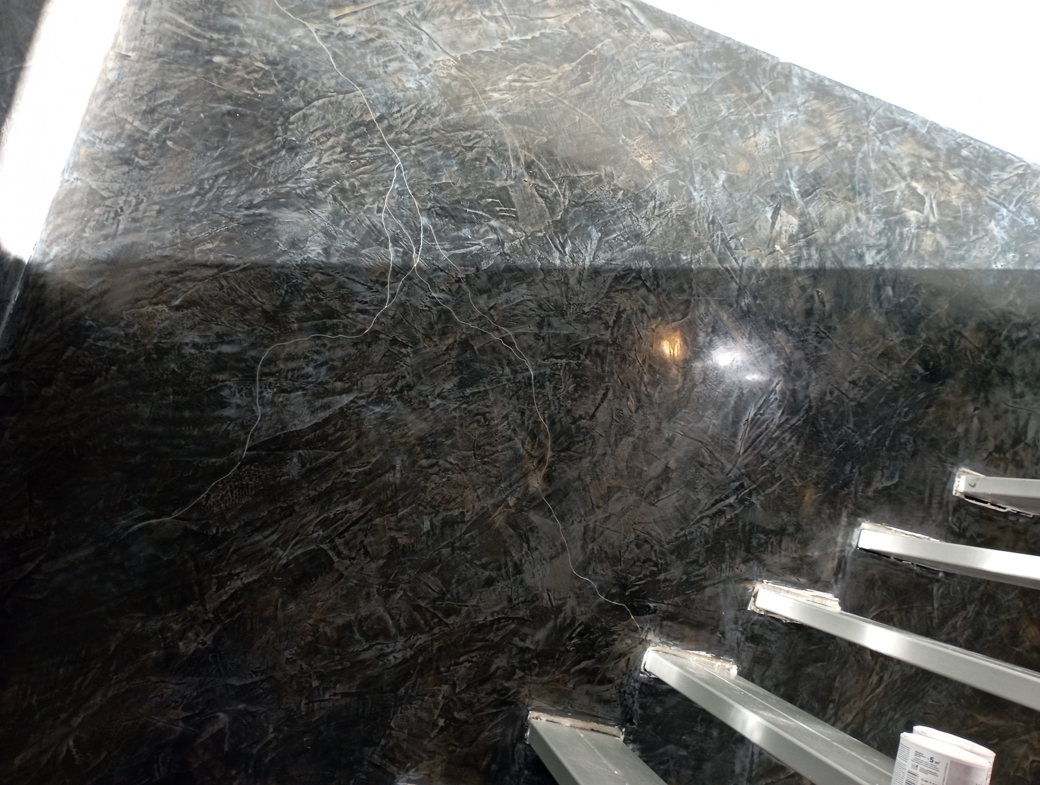 Dekoracyjna ściana w odcieniach szarości z efektem marmuru, widoczna częściowo z metalowymi elementami konstrukcyjnymi schodów. Unikalna faktura i oświetlenie.