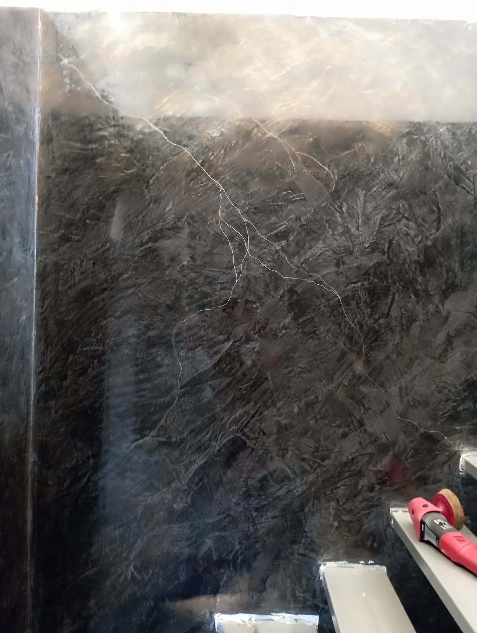 Ściana z efektem dekoracyjnym w odcieniach szarości, imitująca kamień. Widoczne narzędzie do polerowania na schodach. Unikalna faktura i oświetlenie podkreślają głębię.