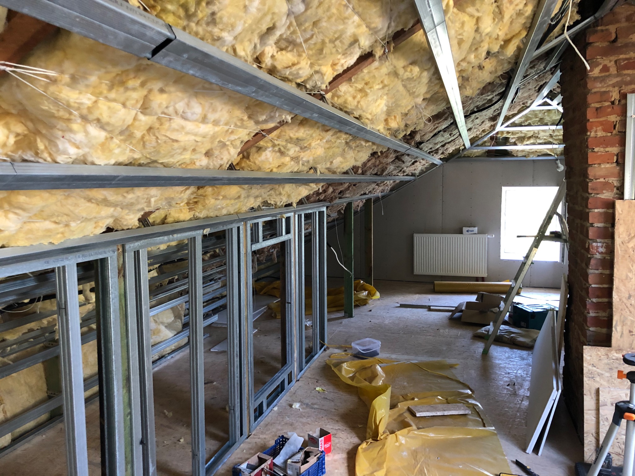 Poddasze w trakcie ocieplania wełną mineralną, widoczne metalowe profile konstrukcyjne ścian działowych, okno w szczycie, rozłożona folia ochronna i narzędzia.