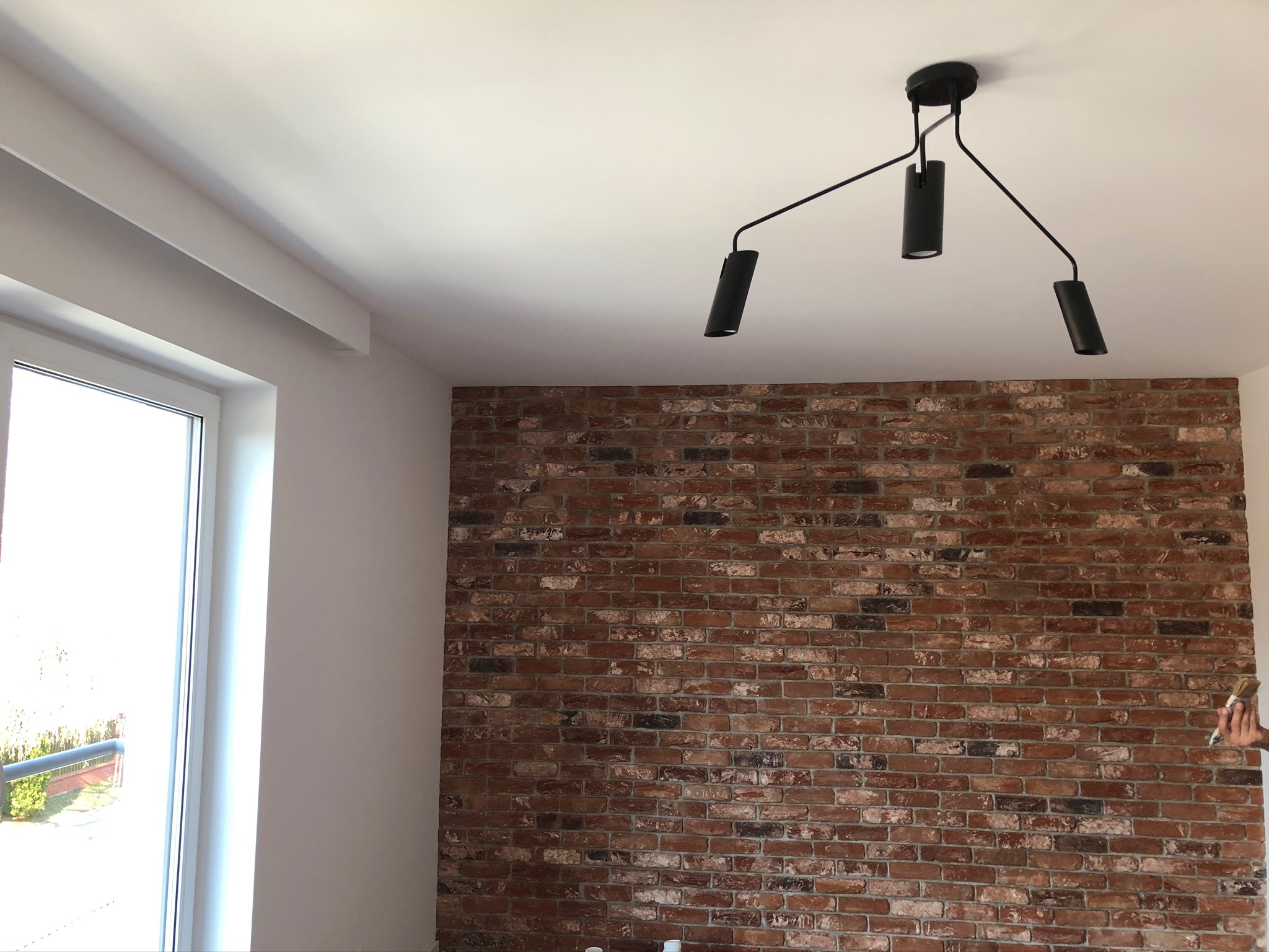 Ściana z cegły klinkierowej w pokoju z nowoczesną lampą sufitową i białym oknem, widoczny fragment dłoni z pędzlem przy ścianie.