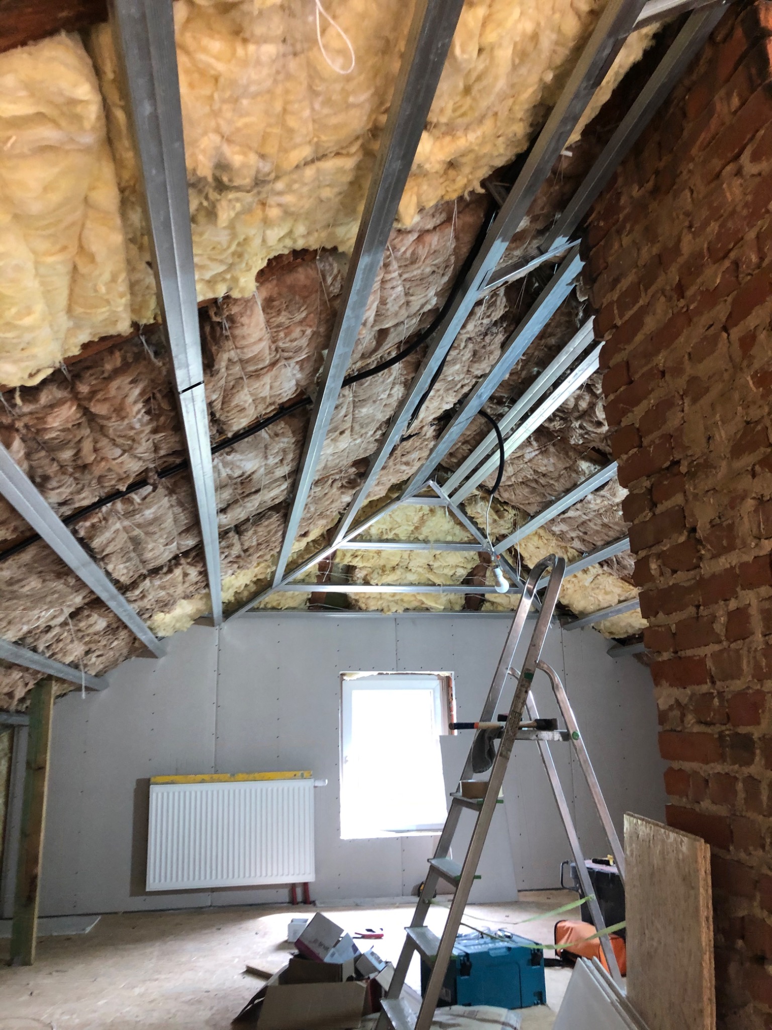 Poddasze w trakcie docieplania wełną mineralną, widoczne metalowe profile konstrukcyjne i fragment murowanej ściany z czerwonej cegły, drabina rozstawiona w pomieszczeniu.