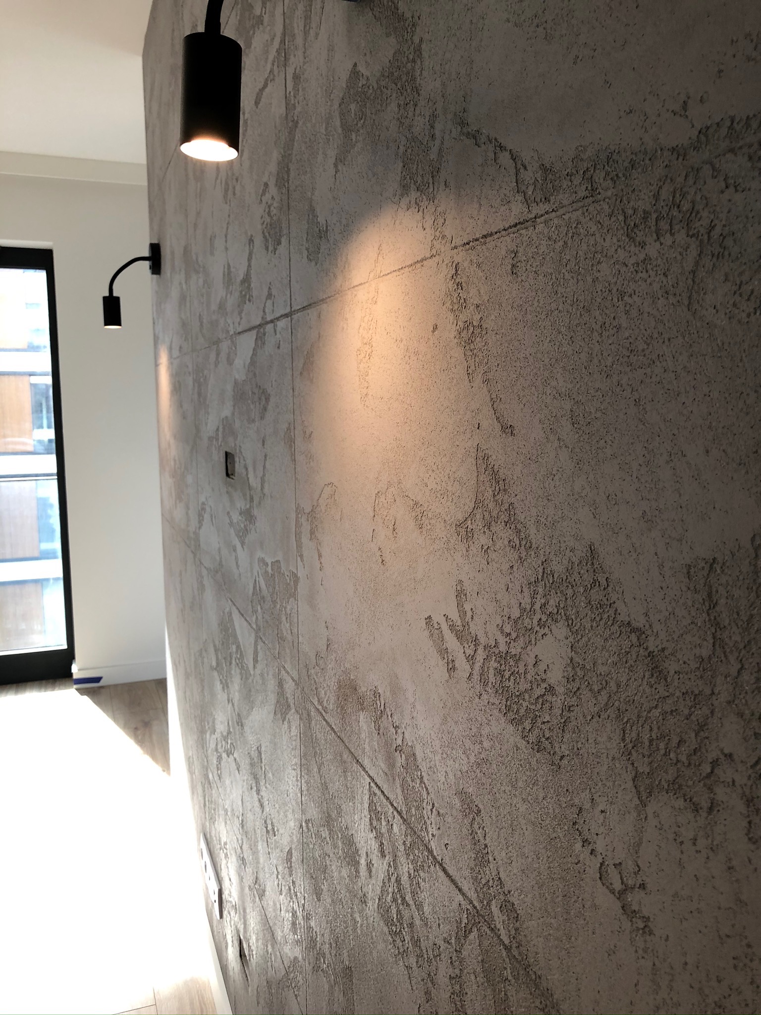 Dekoracyjna ściana z szarego betonu architektonicznego, podświetlona dwoma czarnymi kinkietami, widok z boku.