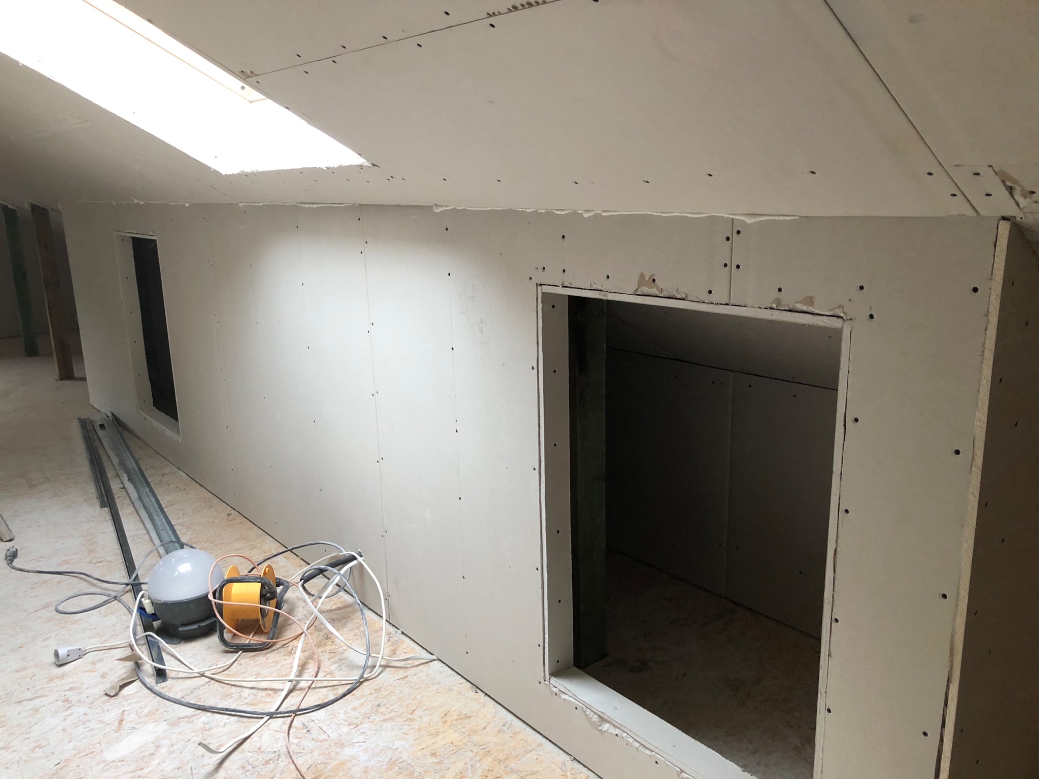 Poddasze z oknem dachowym w trakcie zabudowy płytami gipsowo-kartonowymi, widoczne otwory w ścianach i narzędzia na podłodze.