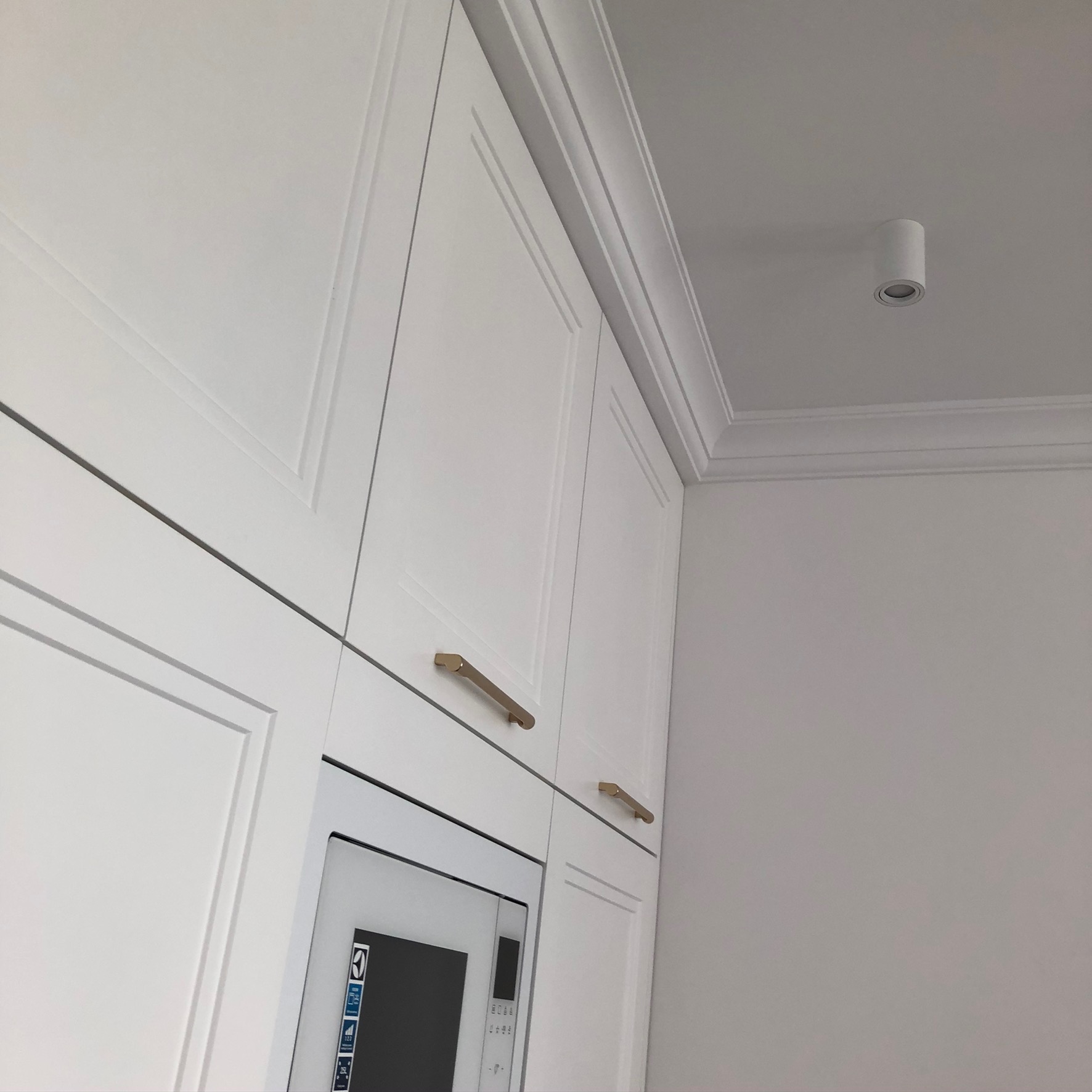 Białe, klasyczne szafki kuchenne z frezowanymi frontami i złotymi uchwytami, zabudowa kuchenna z mikrofalówką, oświetlenie punktowe sufitowe i sztukateria.